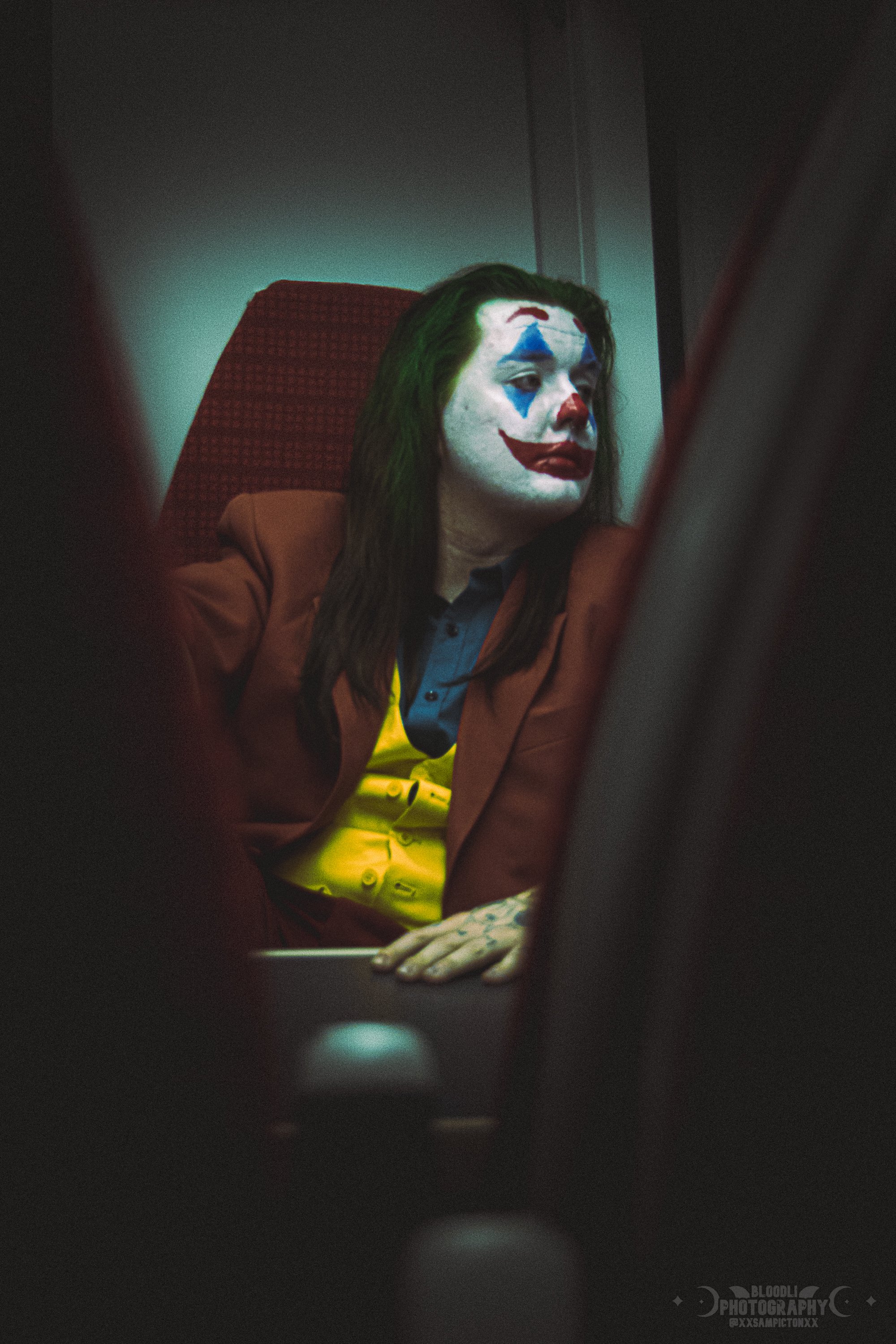 A woman with clown makeup in a clown costume, sitting on a red chair, viewed through a gap in a seat with blurred edges.