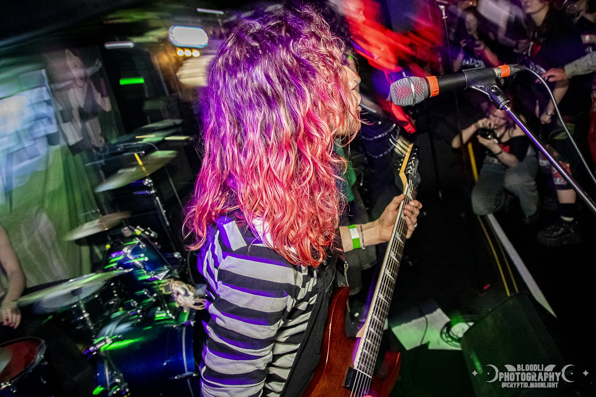 A musician with long, curly pink hair playing an electric guitar and singing into a microphone during a live concert, with colorful stage lighting and an audience in the background.