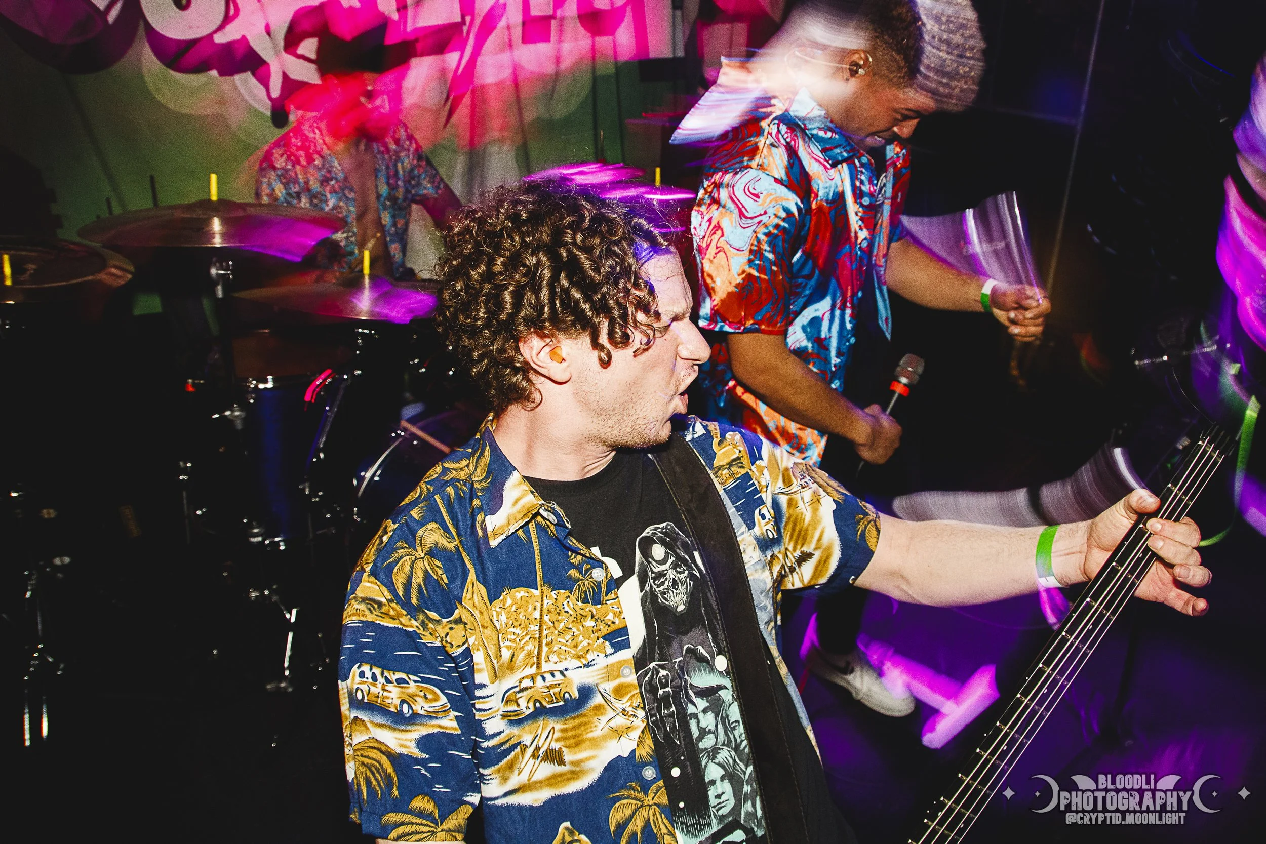 A band performing at a concert with colorful lighting, a drummer in the background, a guitarist playing in the foreground, and two other performers holding microphones.