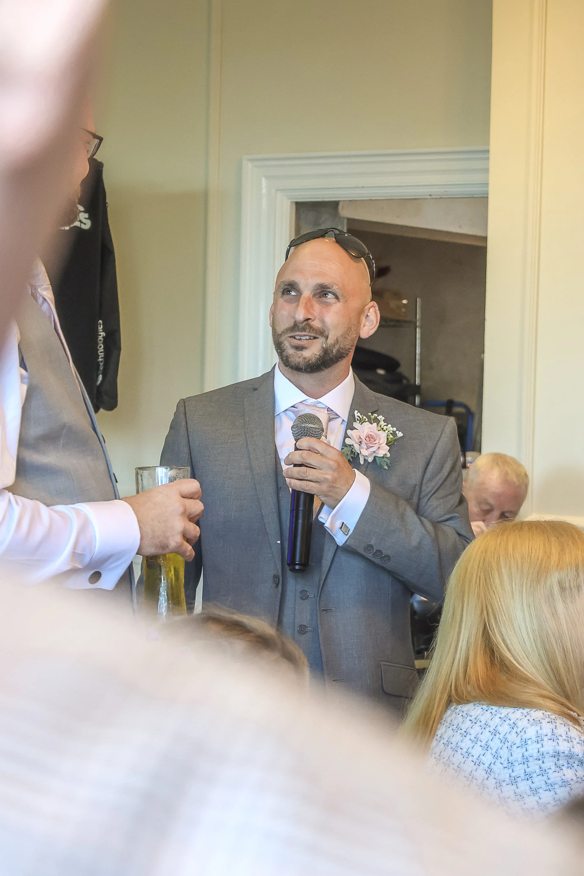 A man in a gray suit holding a microphone and speaking at a formal event with a pink boutonnière on his lapel, while another person in a light-colored jacket holds a drink nearby. Several people are seated or standing in the background in an indoor s