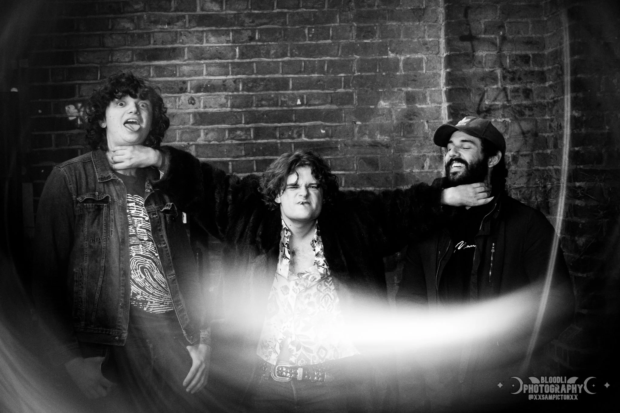 Three young men posing playfully against a brick wall, making funny faces and gestures, in a black and white photo.