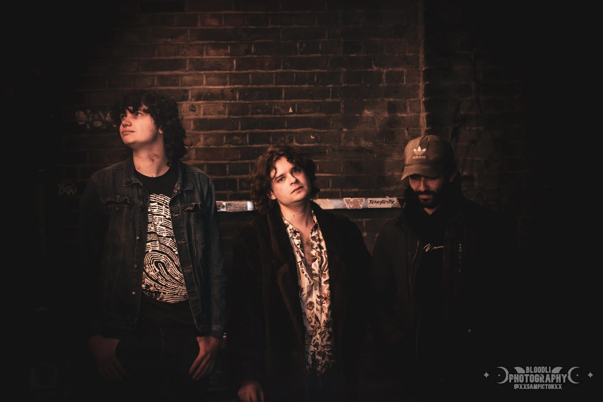 Three young men standing against a brick wall in a dimly lit setting, with the man on the left looking upwards, the man in the middle gazing ahead with a neutral expression, and the man on the right looking downwards.