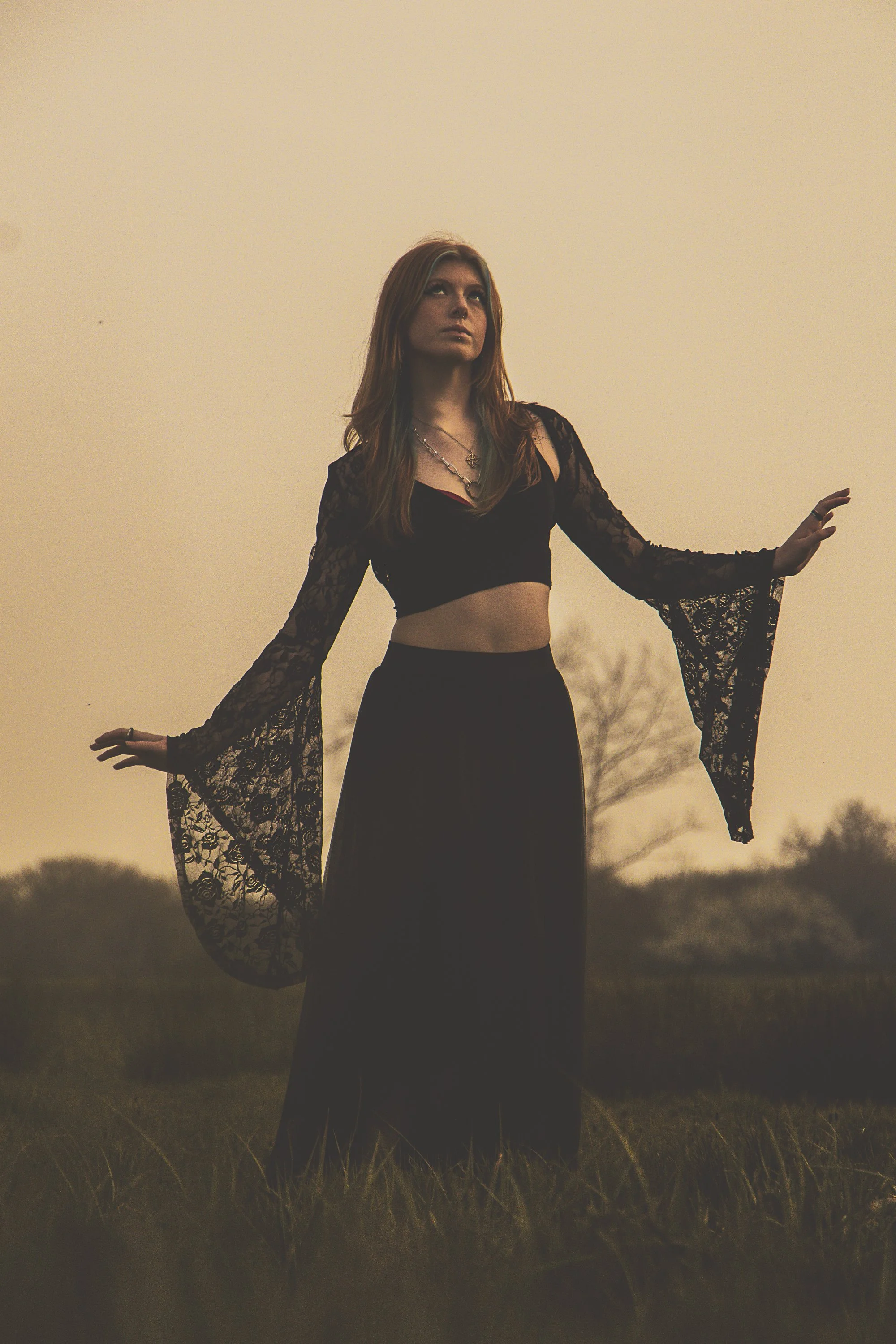 A woman standing outdoors on a grassy field at sunset, wearing a black lace long-sleeve crop top and a long black skirt, with her arms outstretched and looking up.