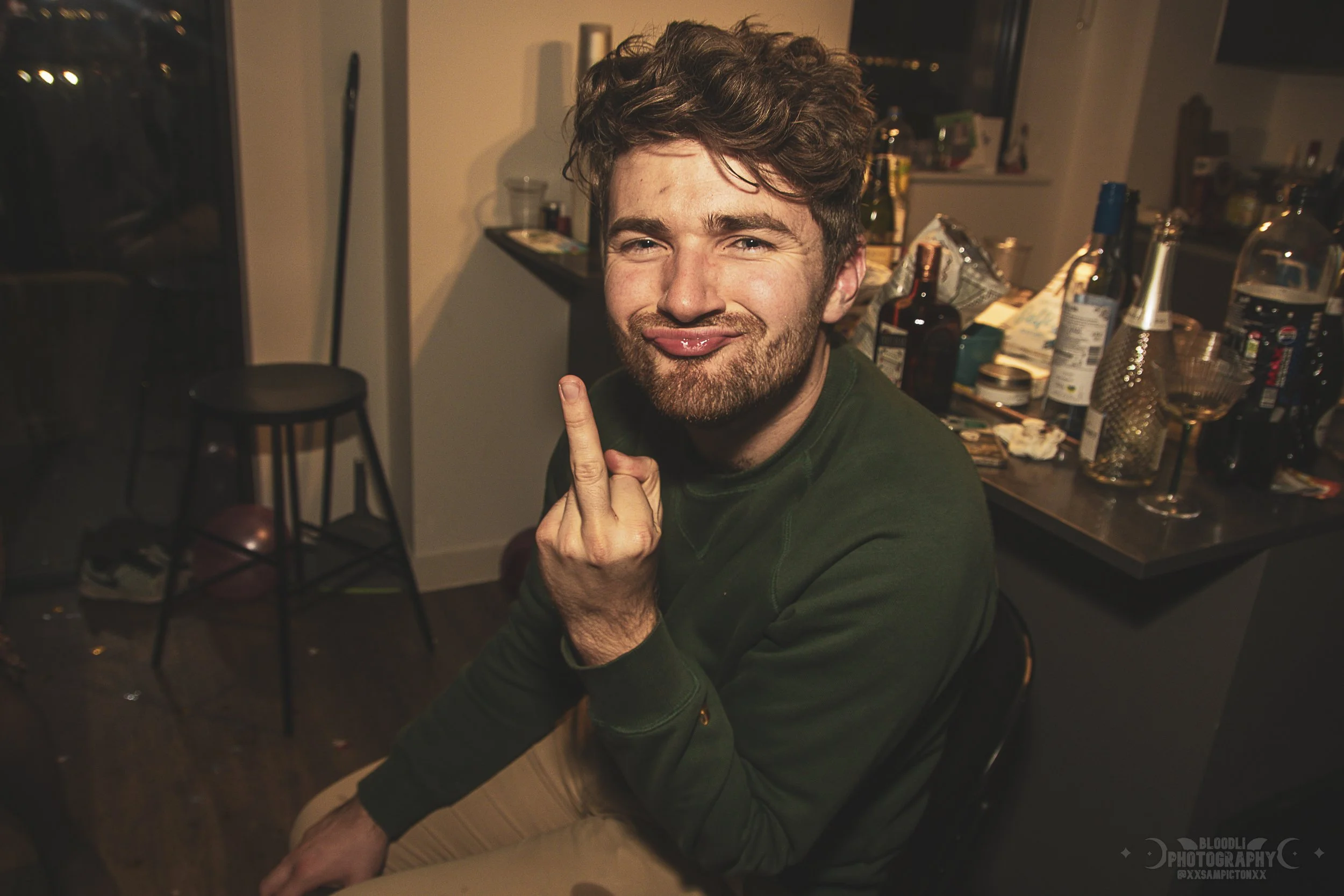 A young man with curly hair and a beard making a silly face, sticking out his tongue and holding up his middle finger, sitting in a cluttered kitchen at night.
