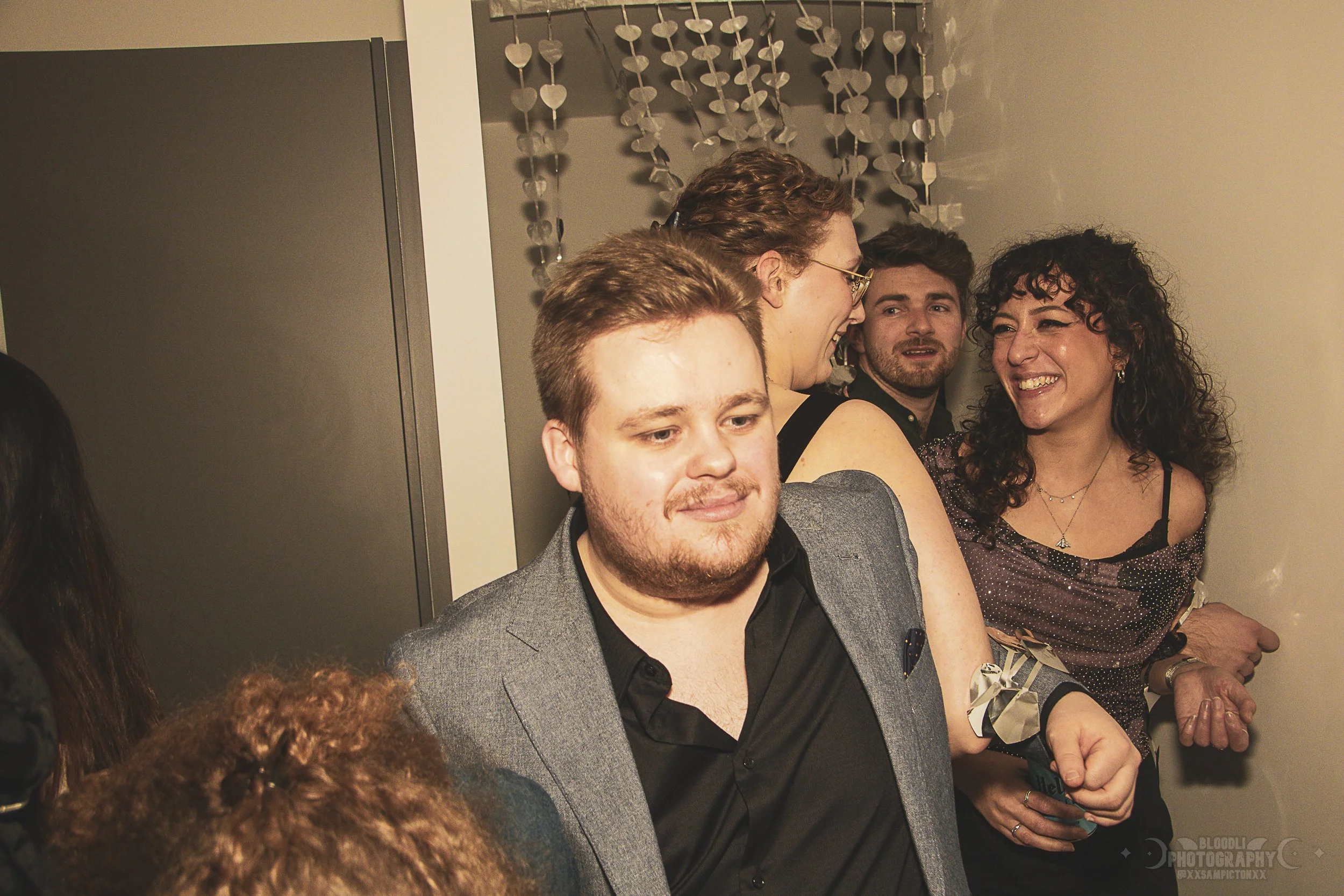 Group of people at a party, engaged in conversation and smiling, standing near a wall decorated with hanging heart-shaped ornaments.