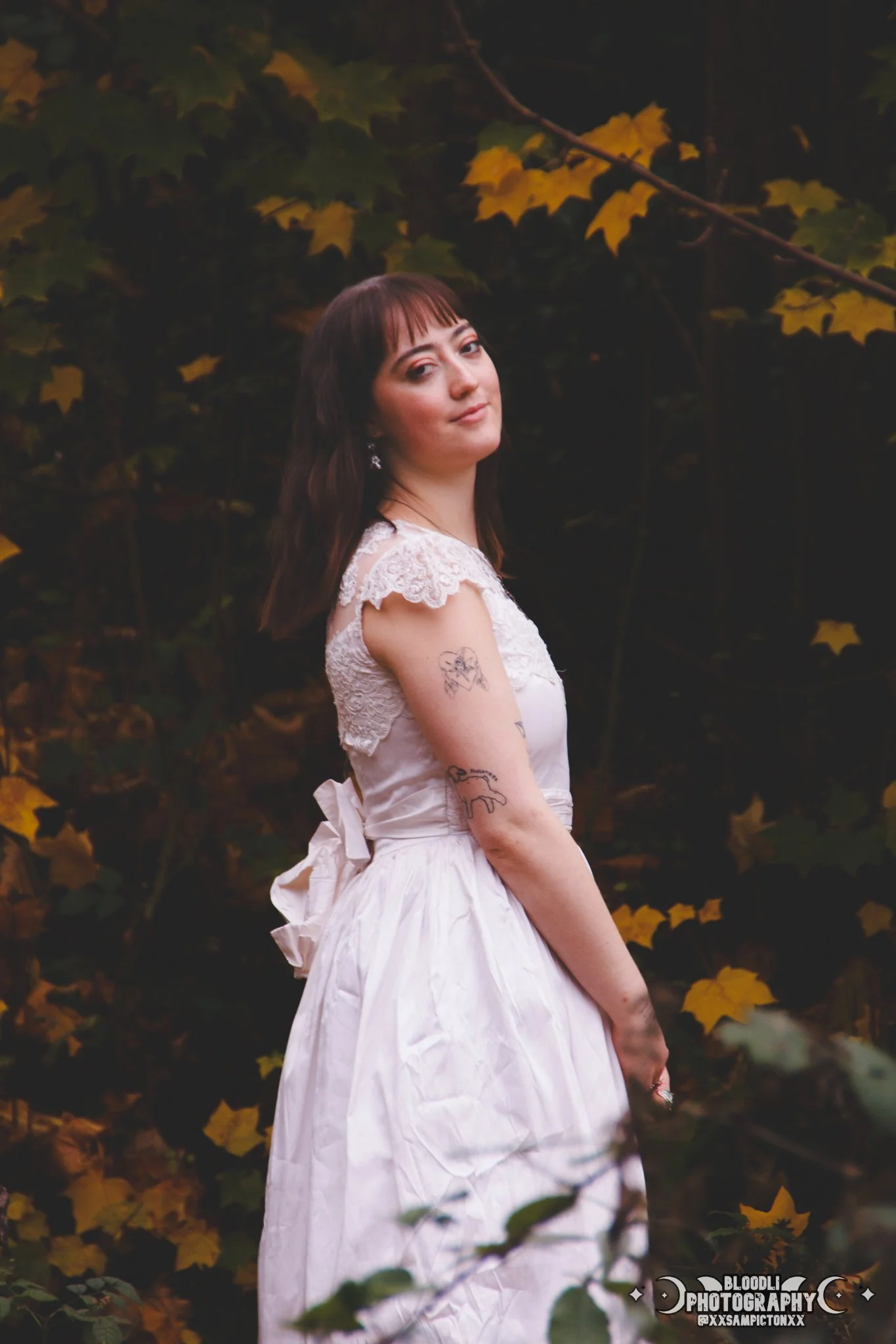 A woman in a white dress with lace details, standing outdoors among yellow leaves and dark foliage, looking at the camera with a slight smile.