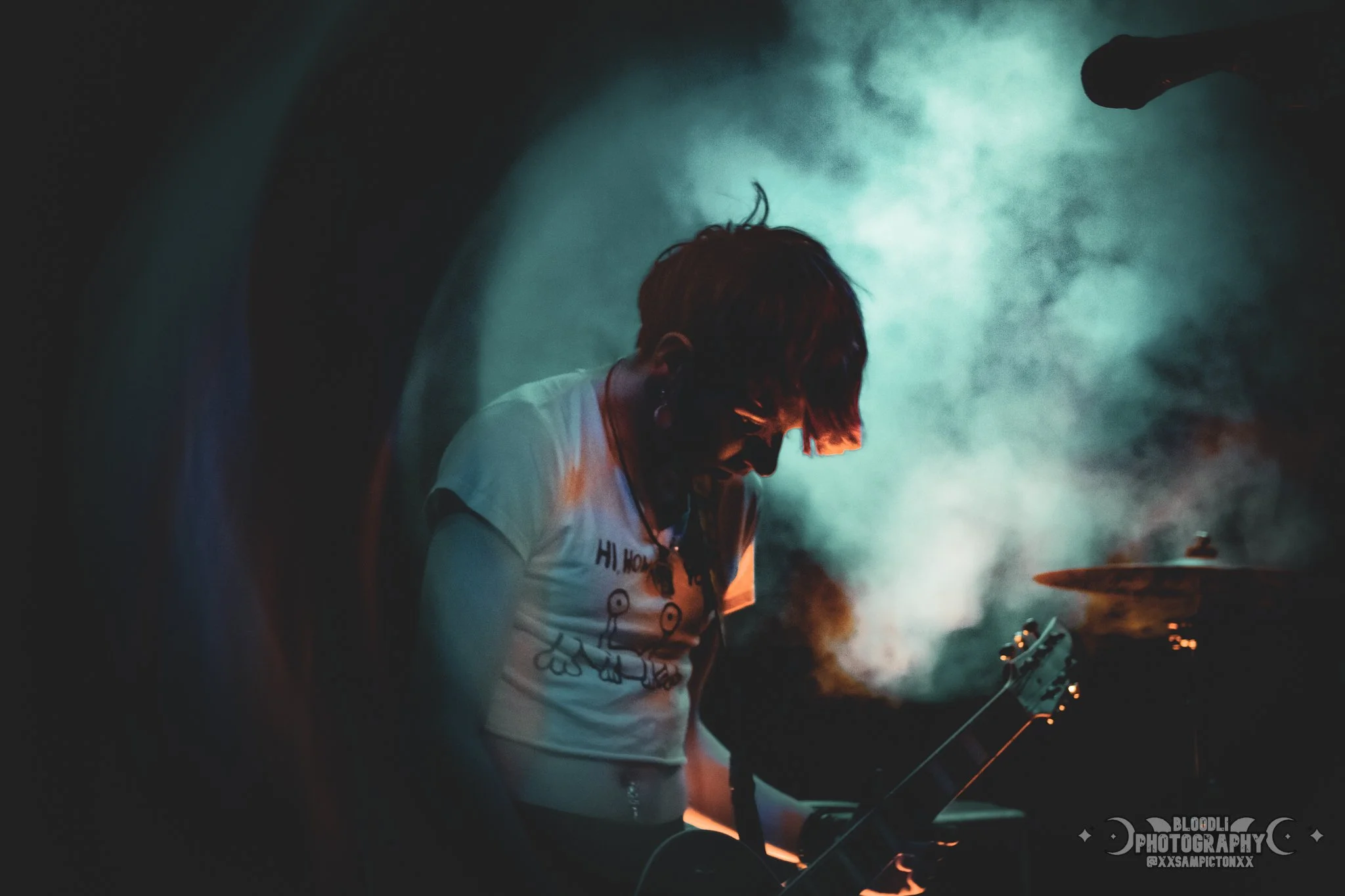 A person with short hair and tattoos, wearing a white t-shirt, playing an electric guitar on stage with blue and orange lighting, surrounded by smoke.