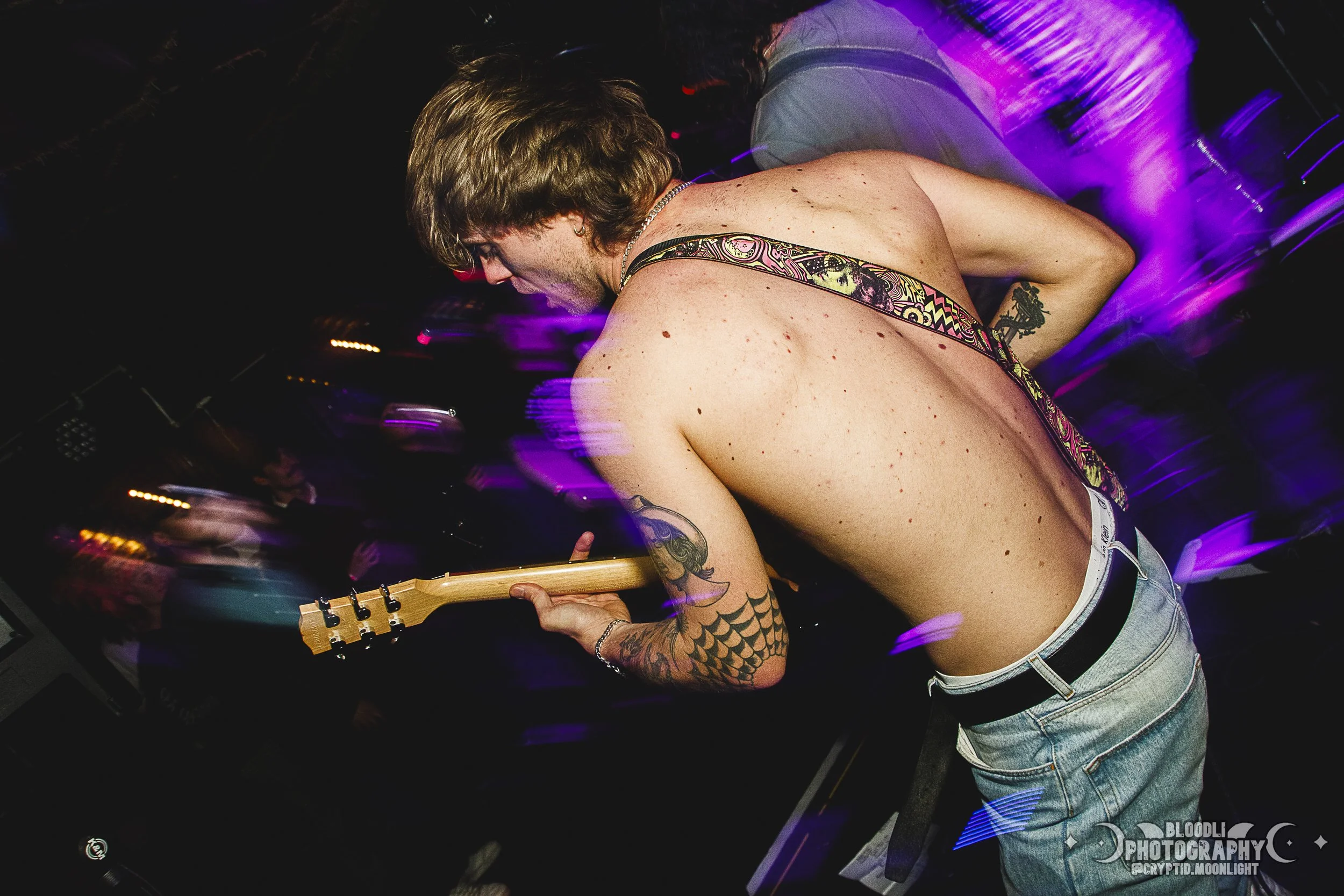 A shirtless man playing an electric guitar at a music event with purple and pink stage lights.