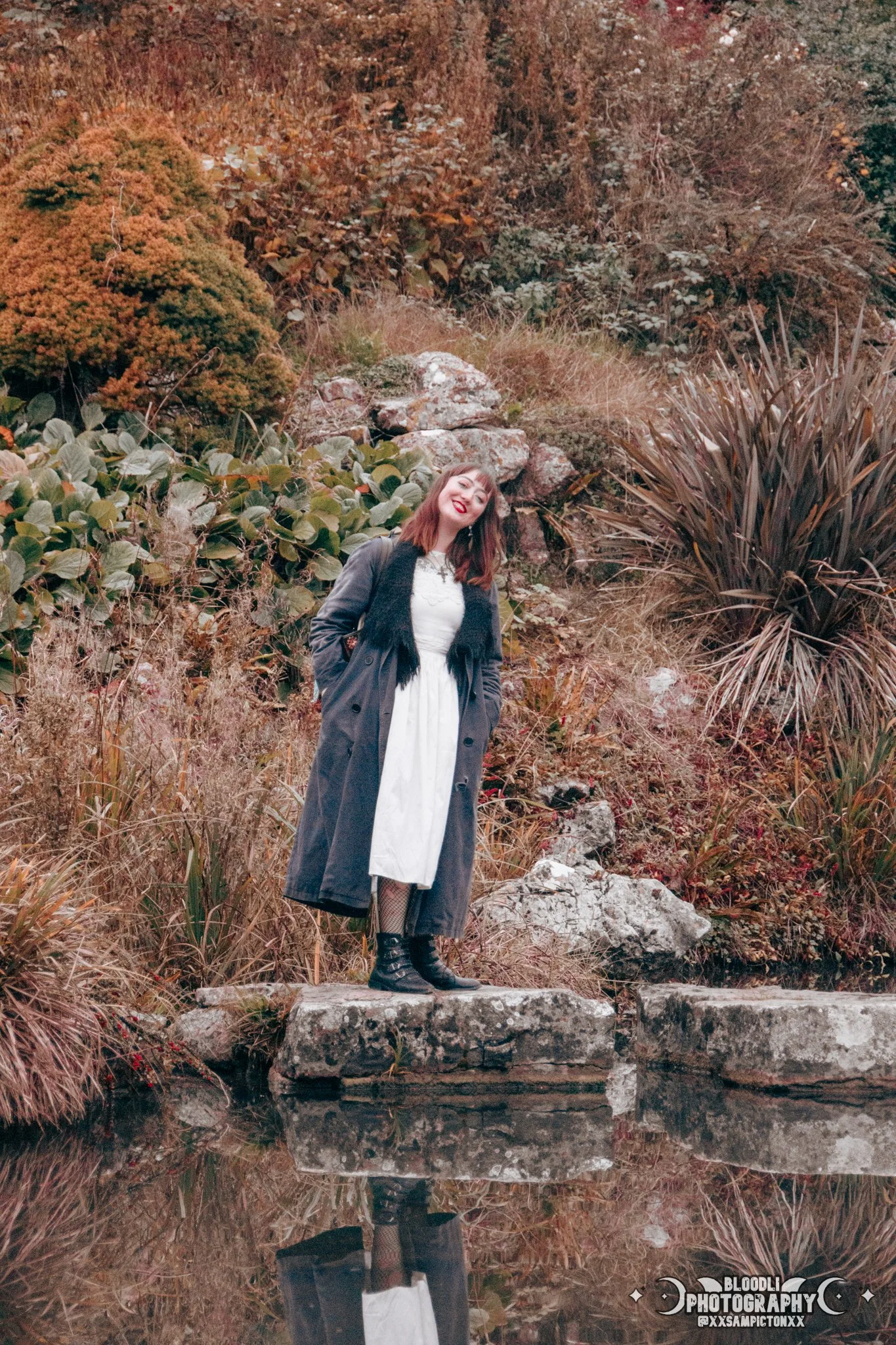 Woman standing on a stone ledge near water with autumn foliage in the background. She is smiling, wearing a white dress, black boots, a long coat, and a scarf.