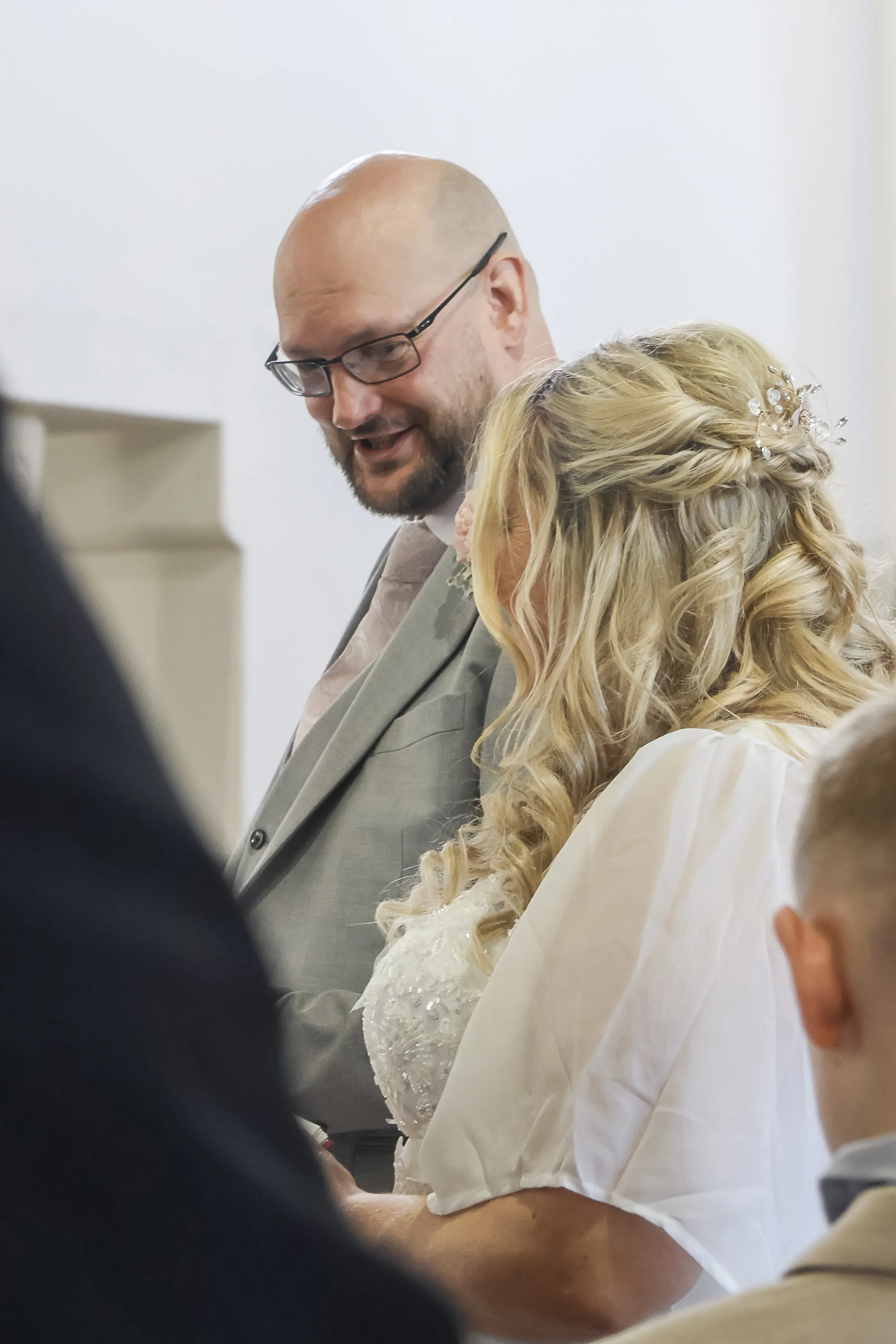 A man with glasses and a beard in a suit is holding hands with a woman with blonde, curled hair decorated with a hairpiece, during a wedding ceremony.