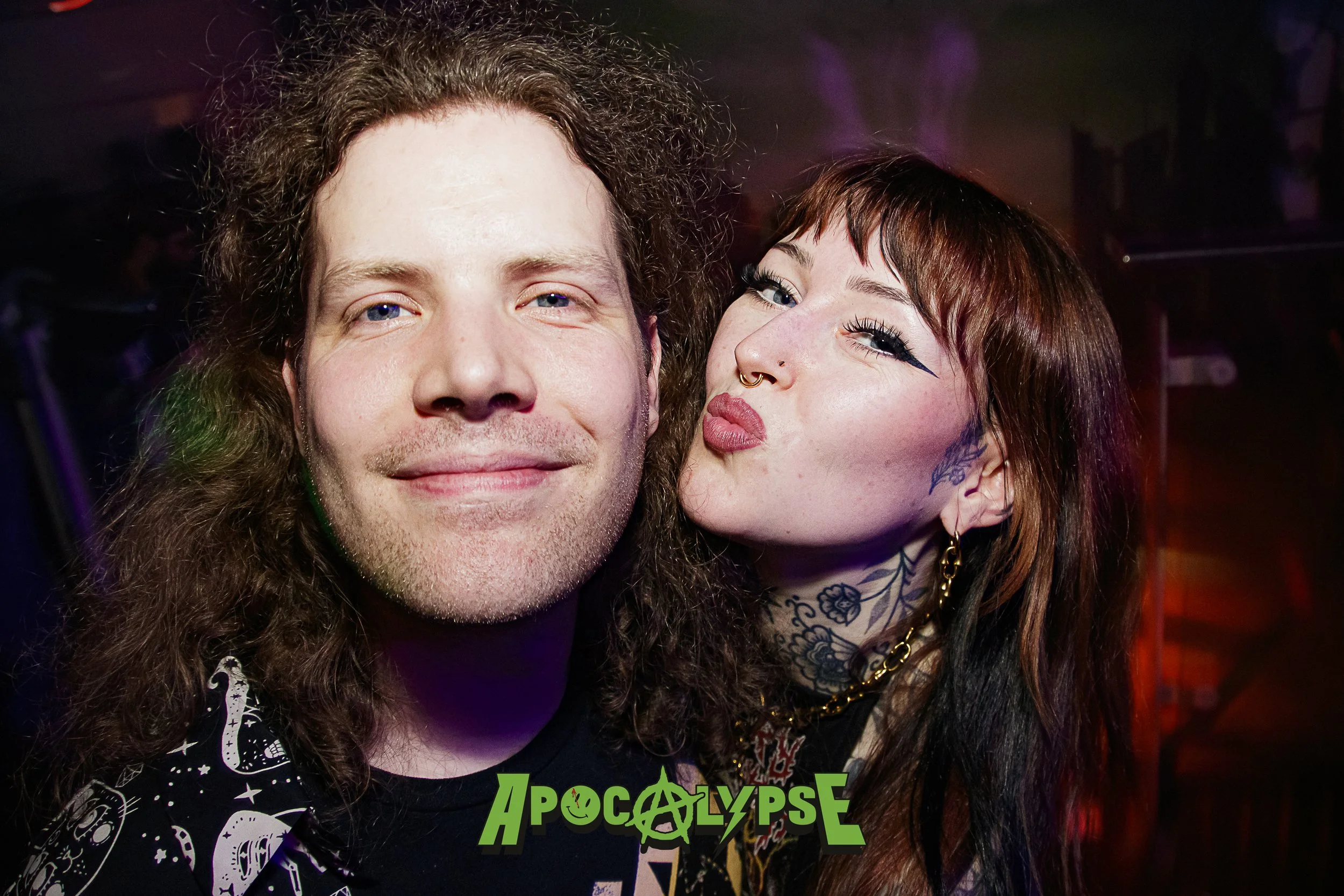 A man with curly brown hair and a woman with tattoos and dark hair making a kissing face at a party, with 