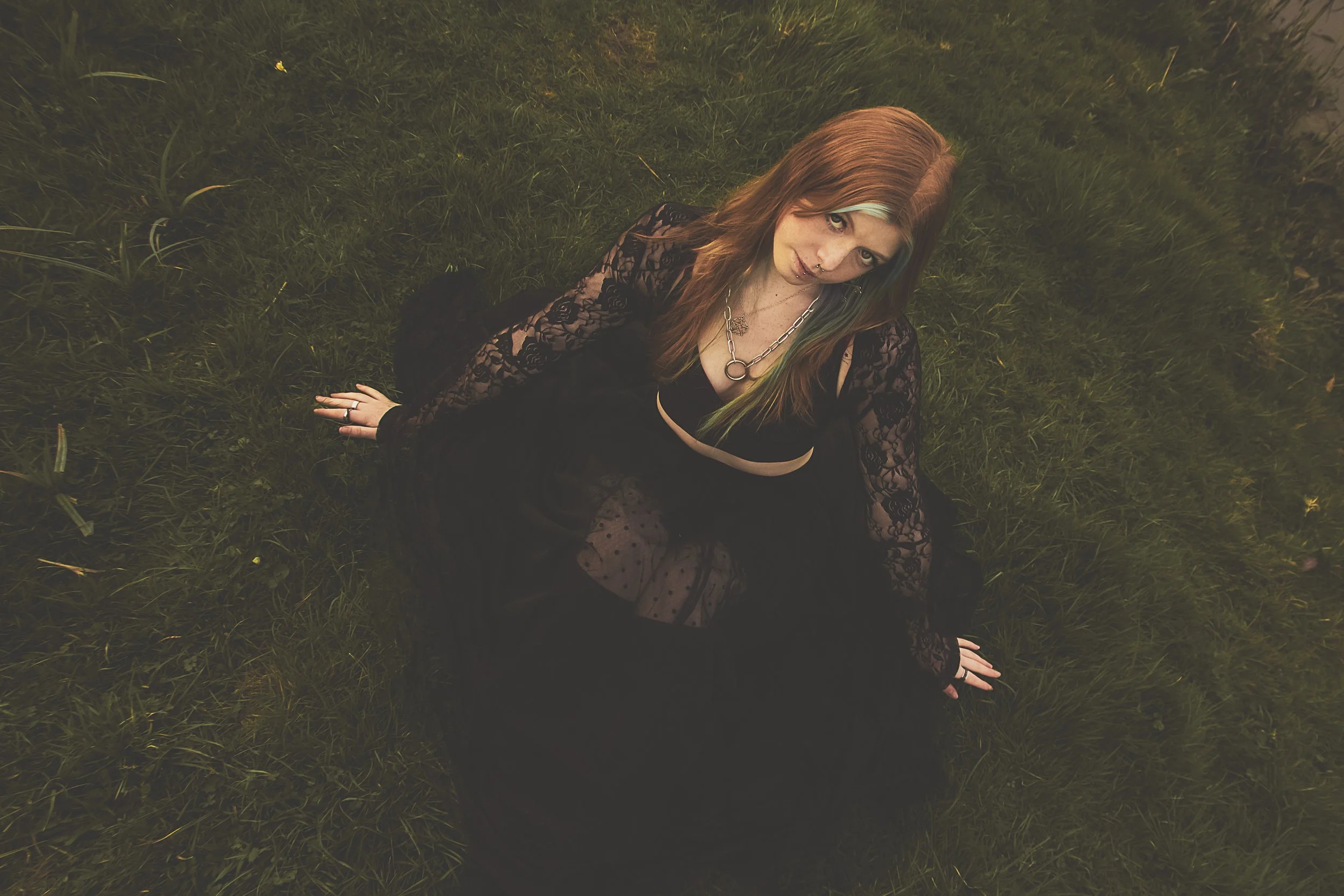 A woman with long reddish-brown hair sits on grass, looking up at the camera. She wears a black lace dress, a black top with a plunging neckline, layered necklaces, and rings on her fingers.