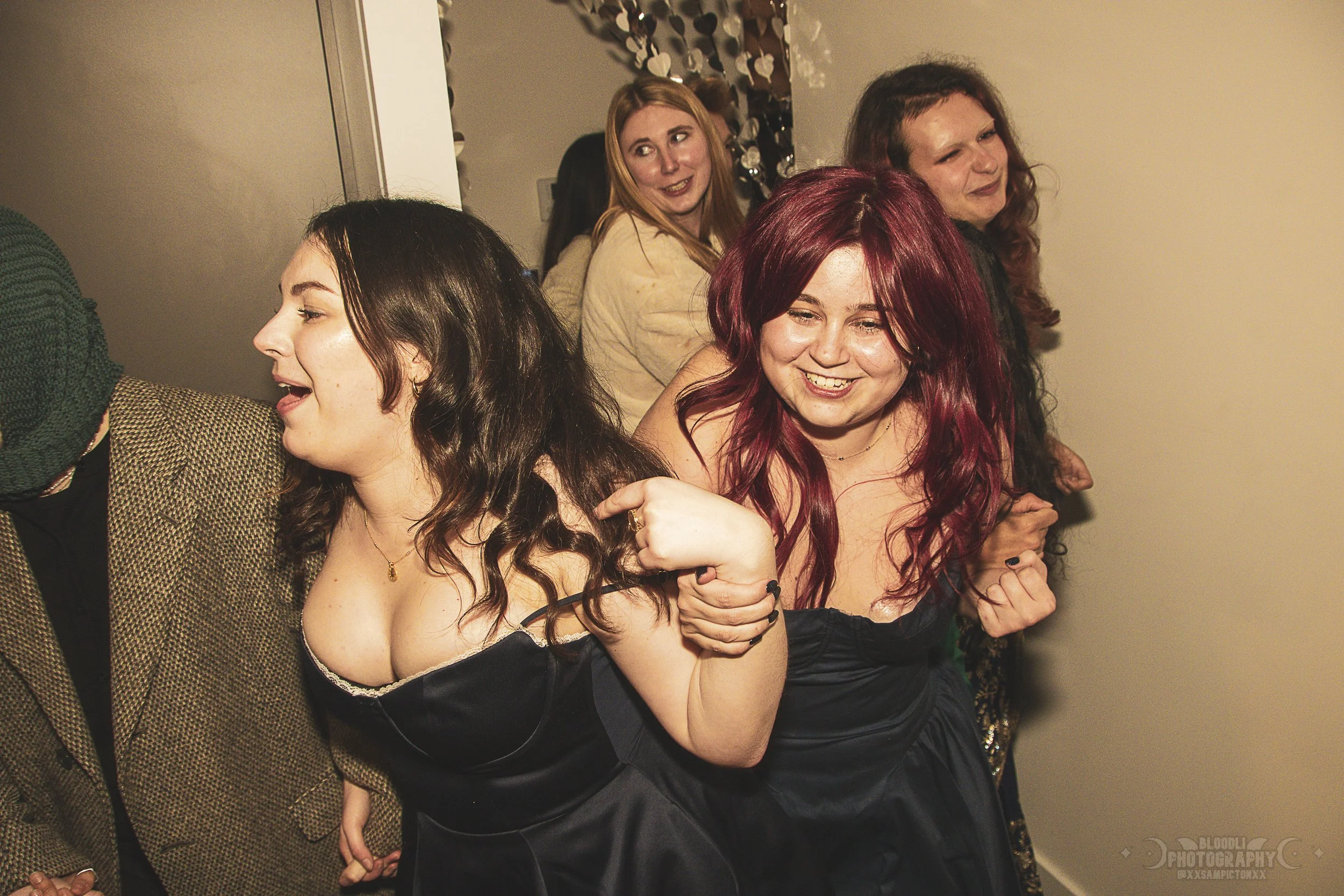 A group of five women smiling and having fun at a party, standing in a line with their arms linked, in an indoor setting.