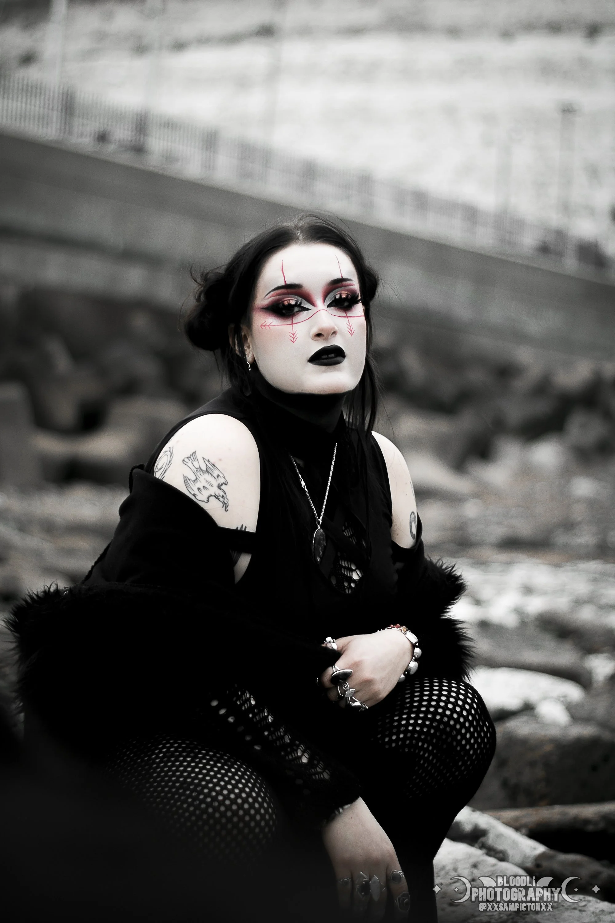 A woman with black hair, dark makeup, and gothic clothing sitting on rocks near a body of water, with a blurred background of a shoreline and a fence.