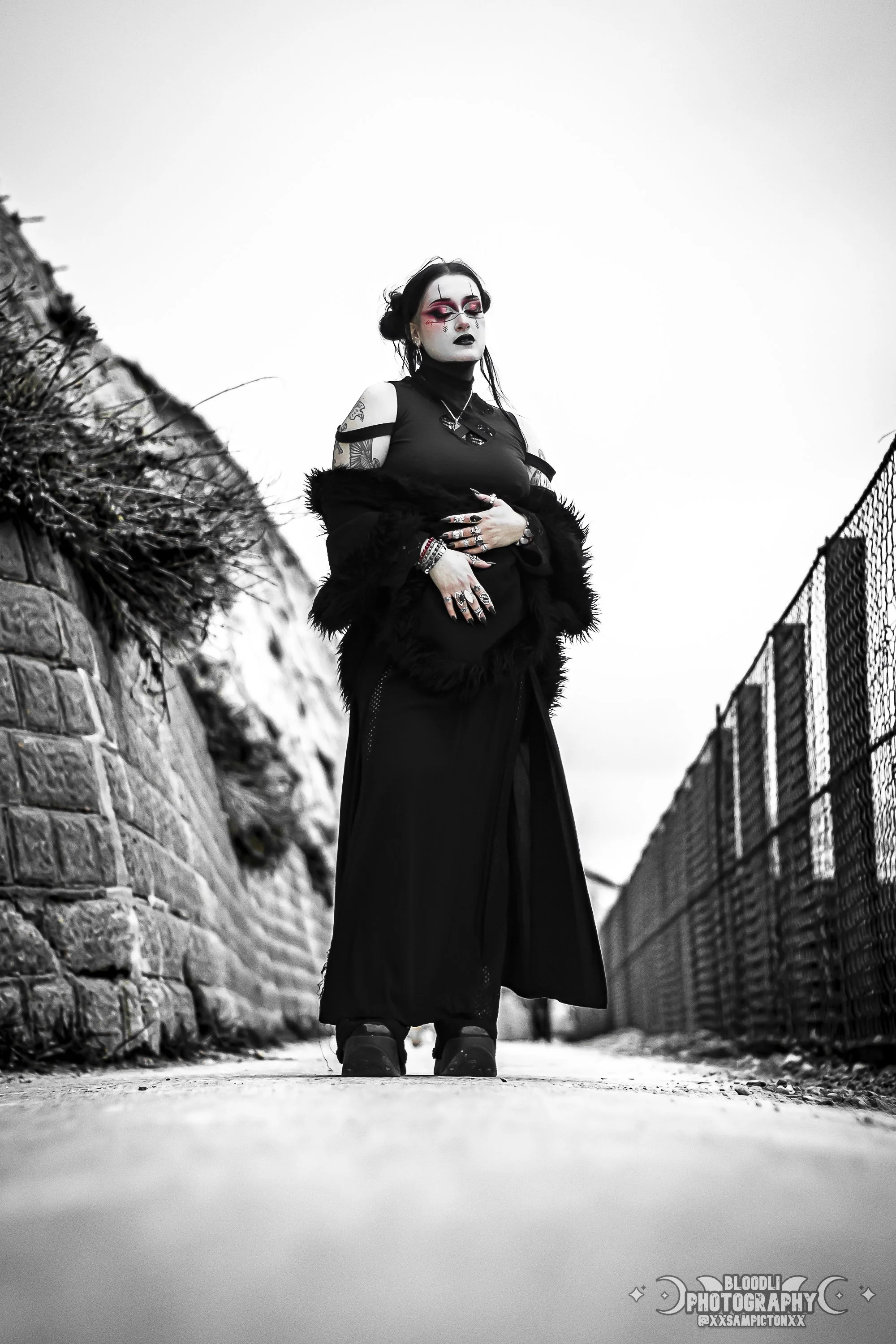 A woman dressed in gothic style clothing stands on a pathway between a stone wall and a chain-link fence, with a cloudy sky in the background.