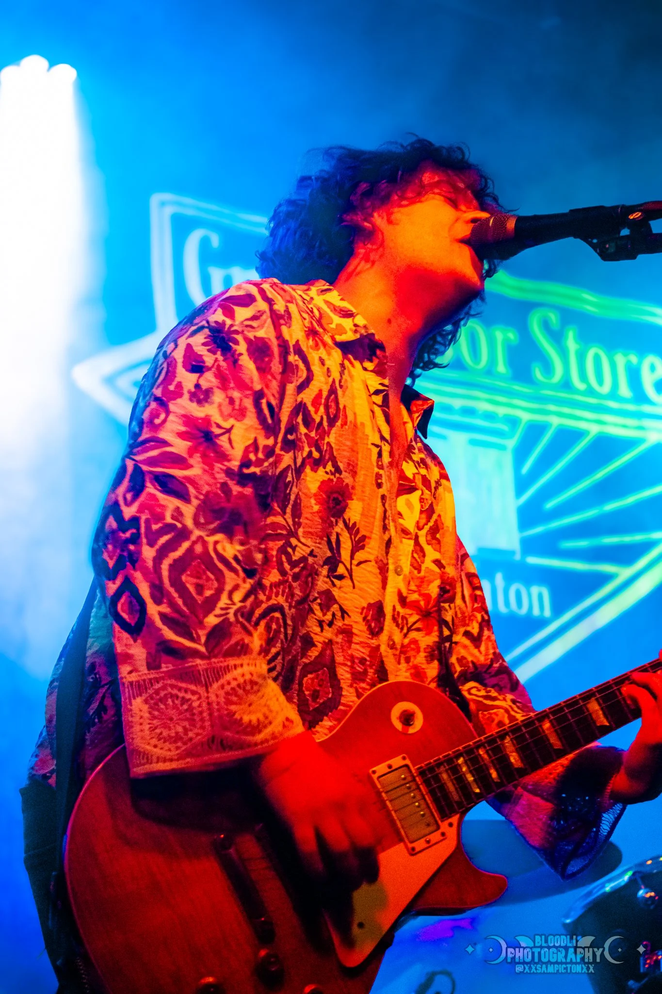 Musician with curly dark hair wearing a colorful, patterned shirt playing electric guitar and singing into a microphone on stage with neon sign in background.