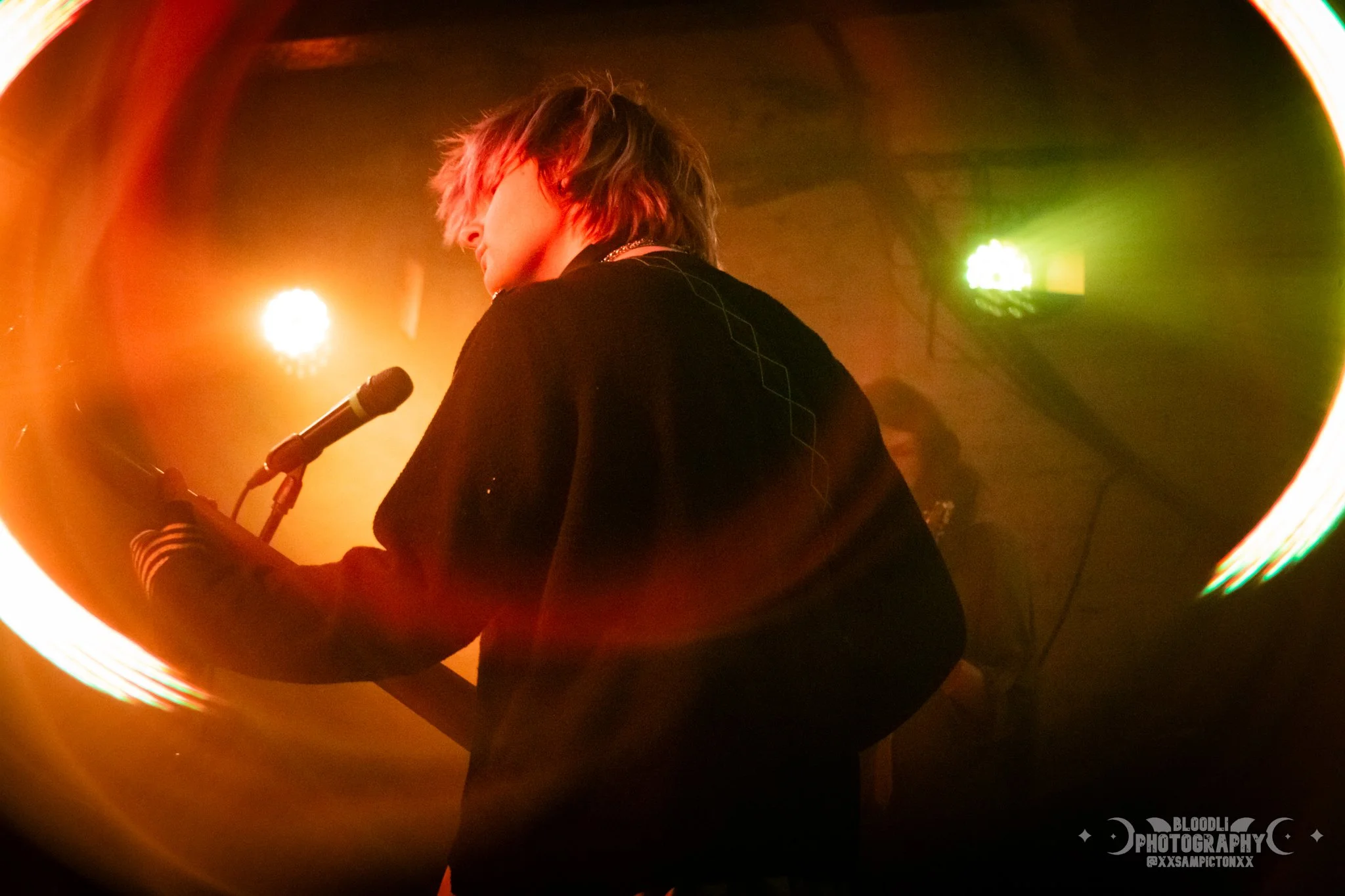 A person with blond hair and black clothing stands on stage with a microphone in front of them, illuminated by colorful stage lights in orange, green, and yellow, creating a vibrant atmosphere.