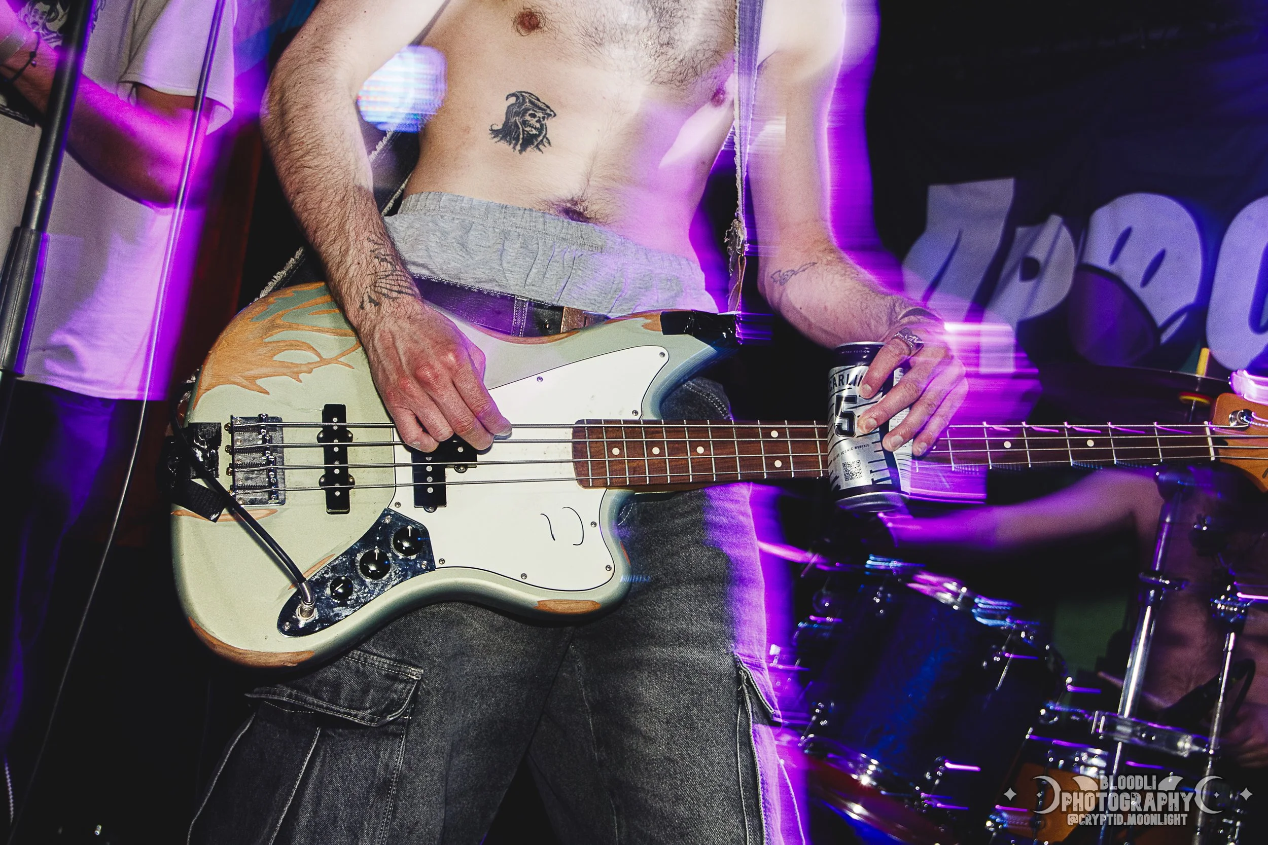 A shirtless man with tattoos playing an electric bass guitar at a music event, with purple and pink stage lights in the background.