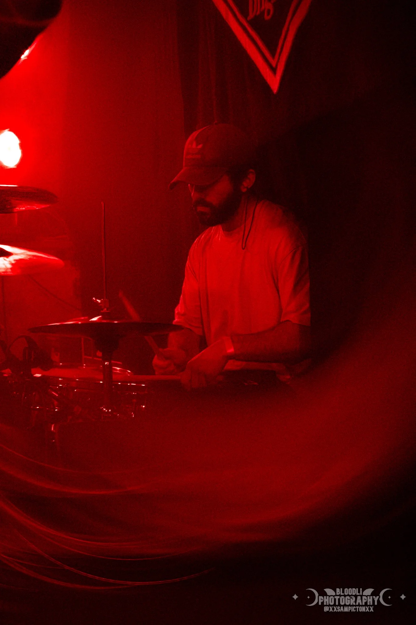 A man wearing a baseball cap and glasses is playing a drum kit in a dimly lit room with red lighting.