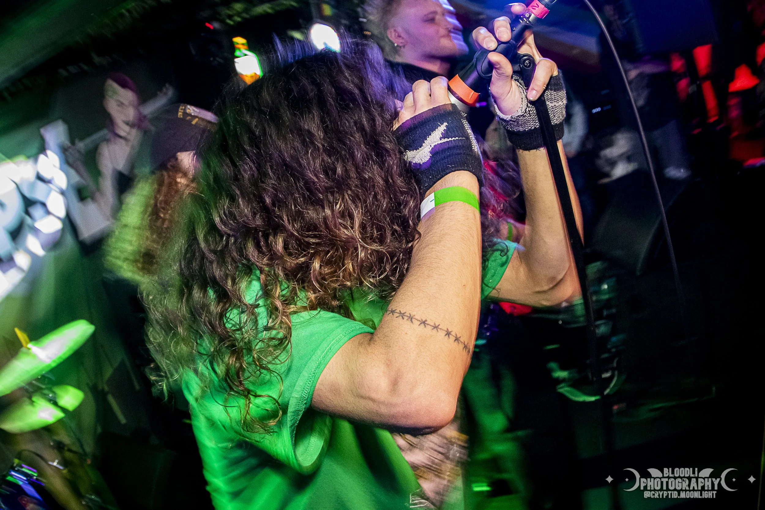 A person with curly hair singing into a microphone on stage, wearing a green shirt, with colorful lights in the background.