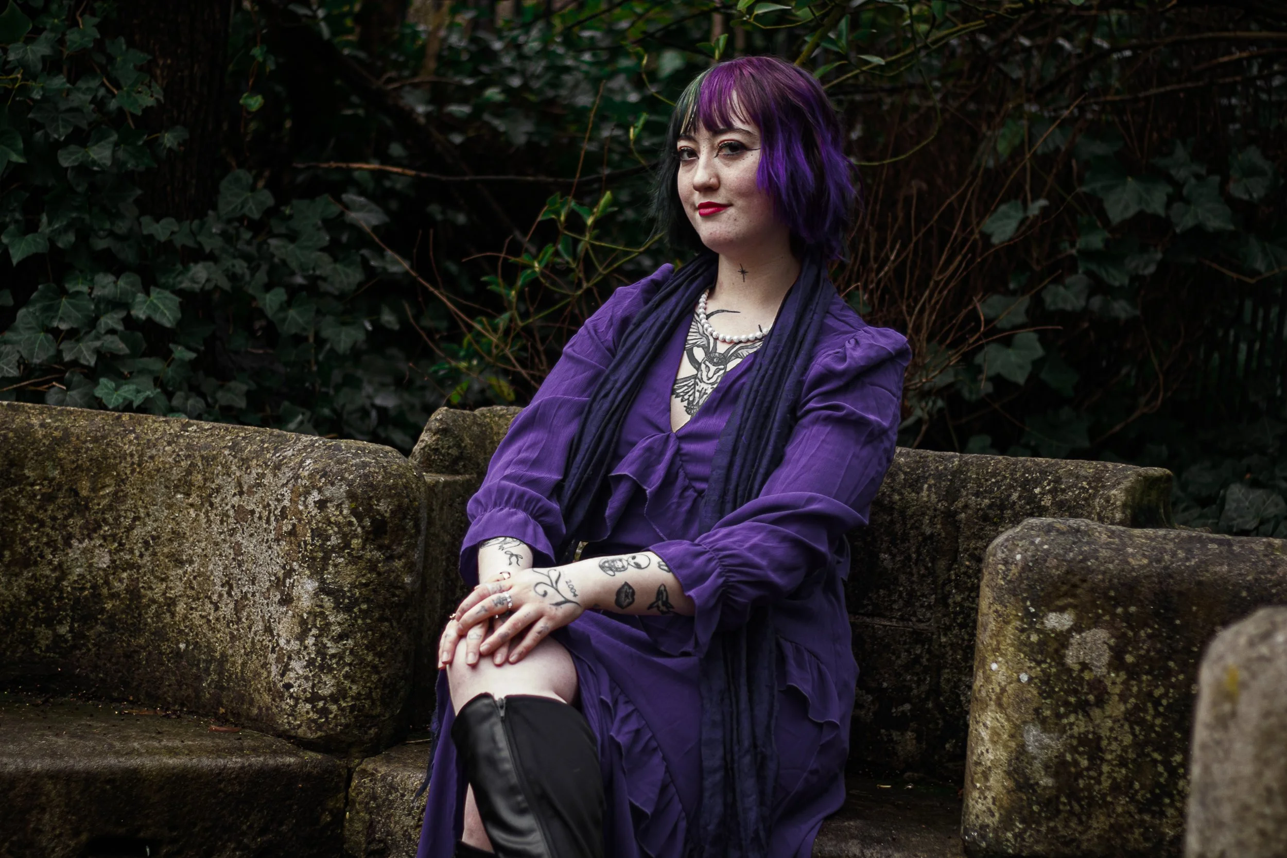 A young woman with black and purple hair sitting on a mossy stone bench in a garden, wearing a purple dress, black scarf, and thigh-high black boots, with tattoos on her arms and chest.