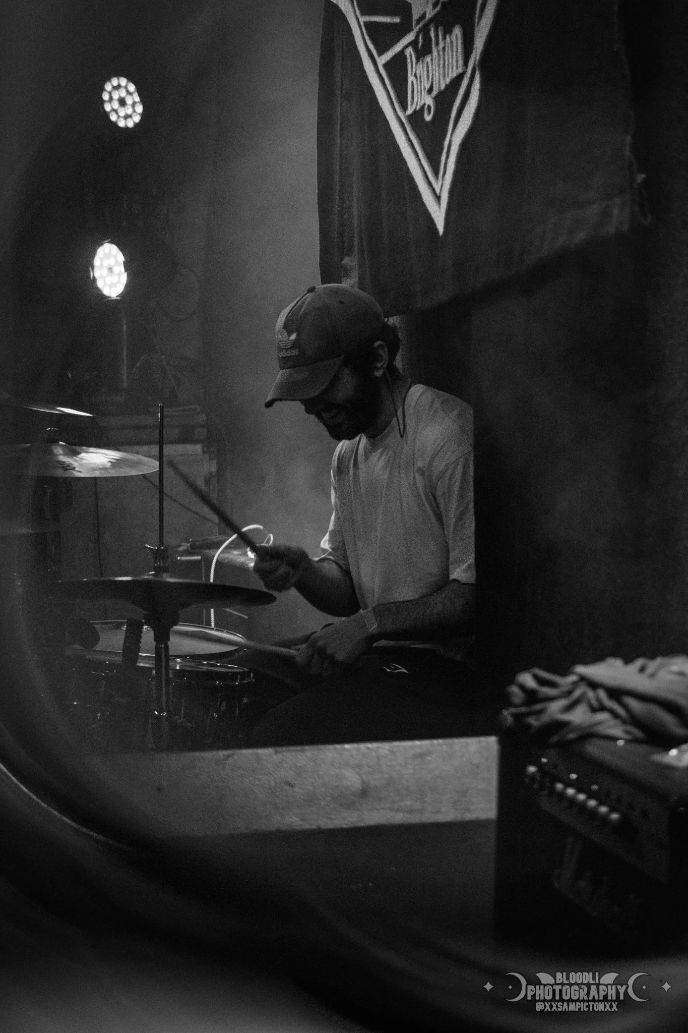 A person wearing a baseball cap playing drums in a dark, smoky environment, possibly a music venue or stage.