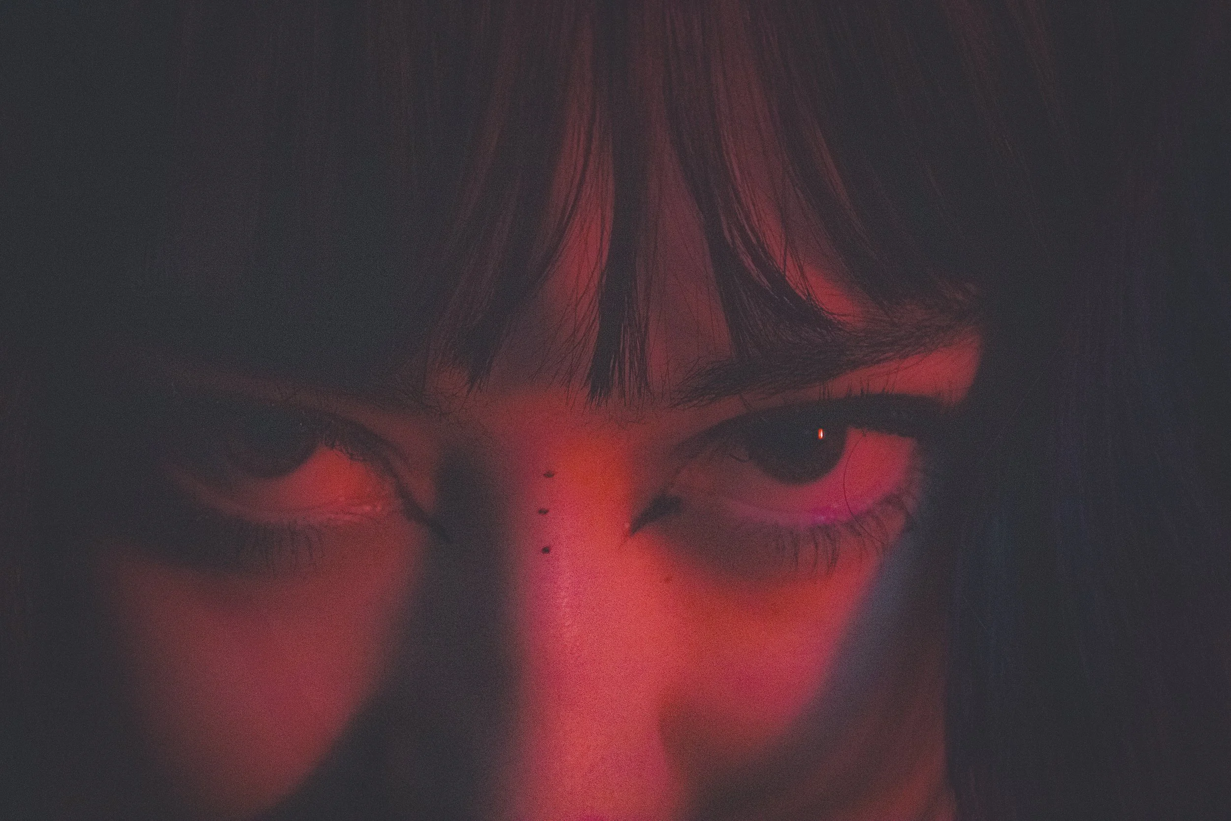 Close-up of a person's face with intense eyes, black hair with bangs, illuminated by red lighting.
