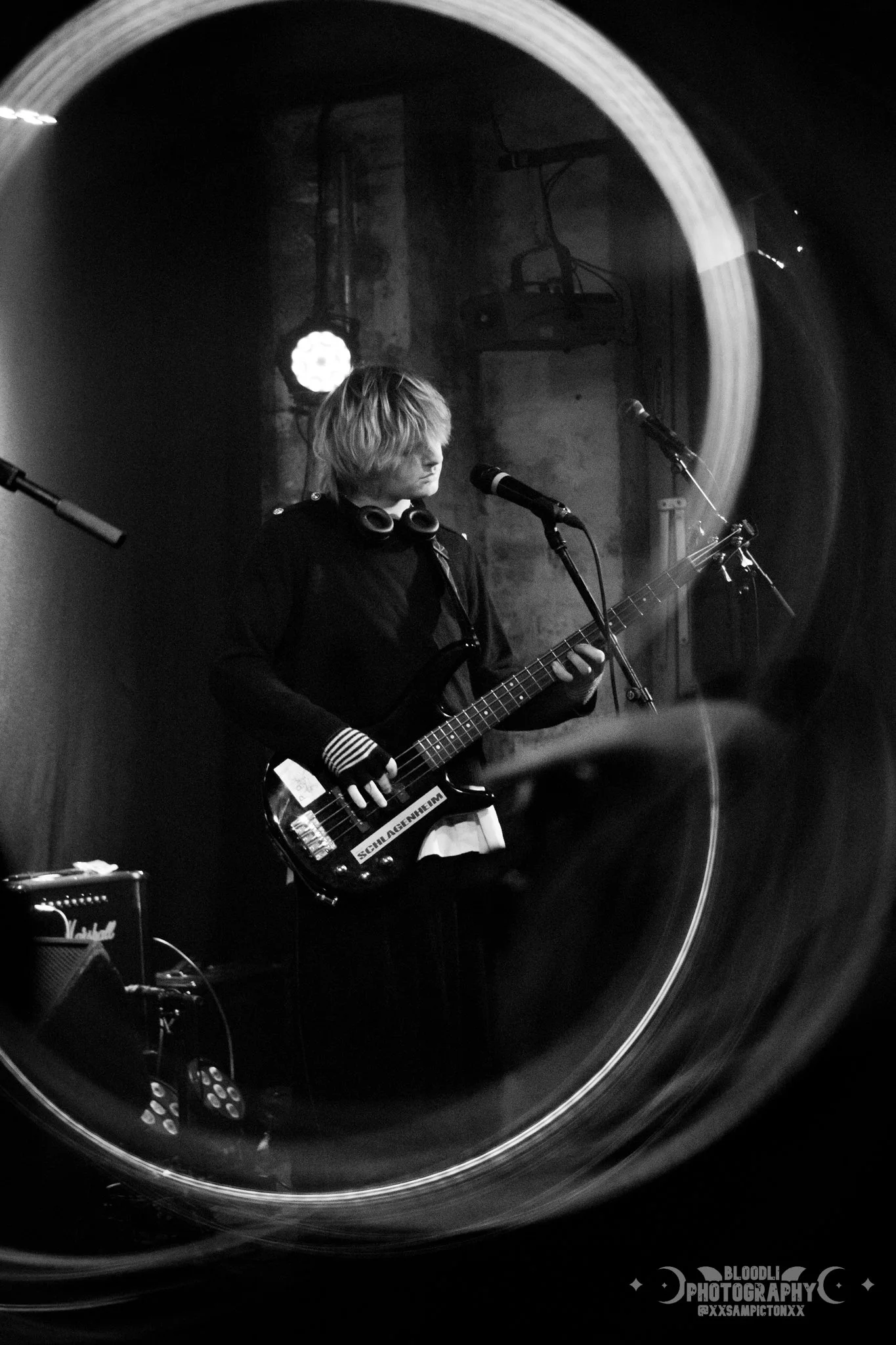A black-and-white photo of a young person with blonde hair playing an electric guitar on stage, with microphones and stage equipment around, seen through a circular reflection.