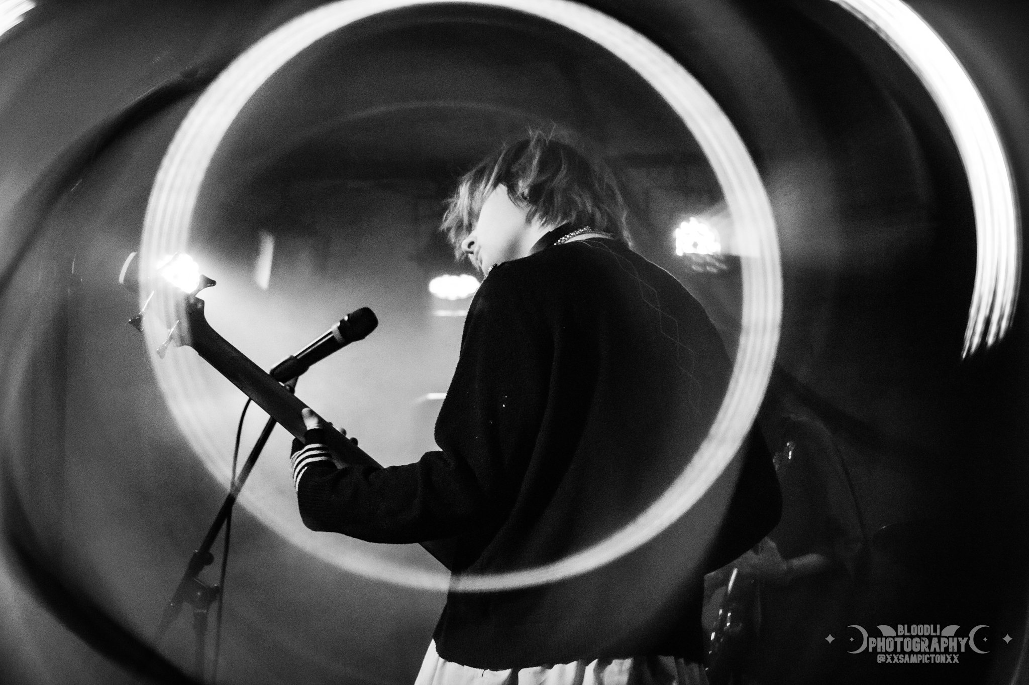 A woman with short hair singing or playing guitar on stage, shot through a circular object creating a ring around her, with blurred background lights.