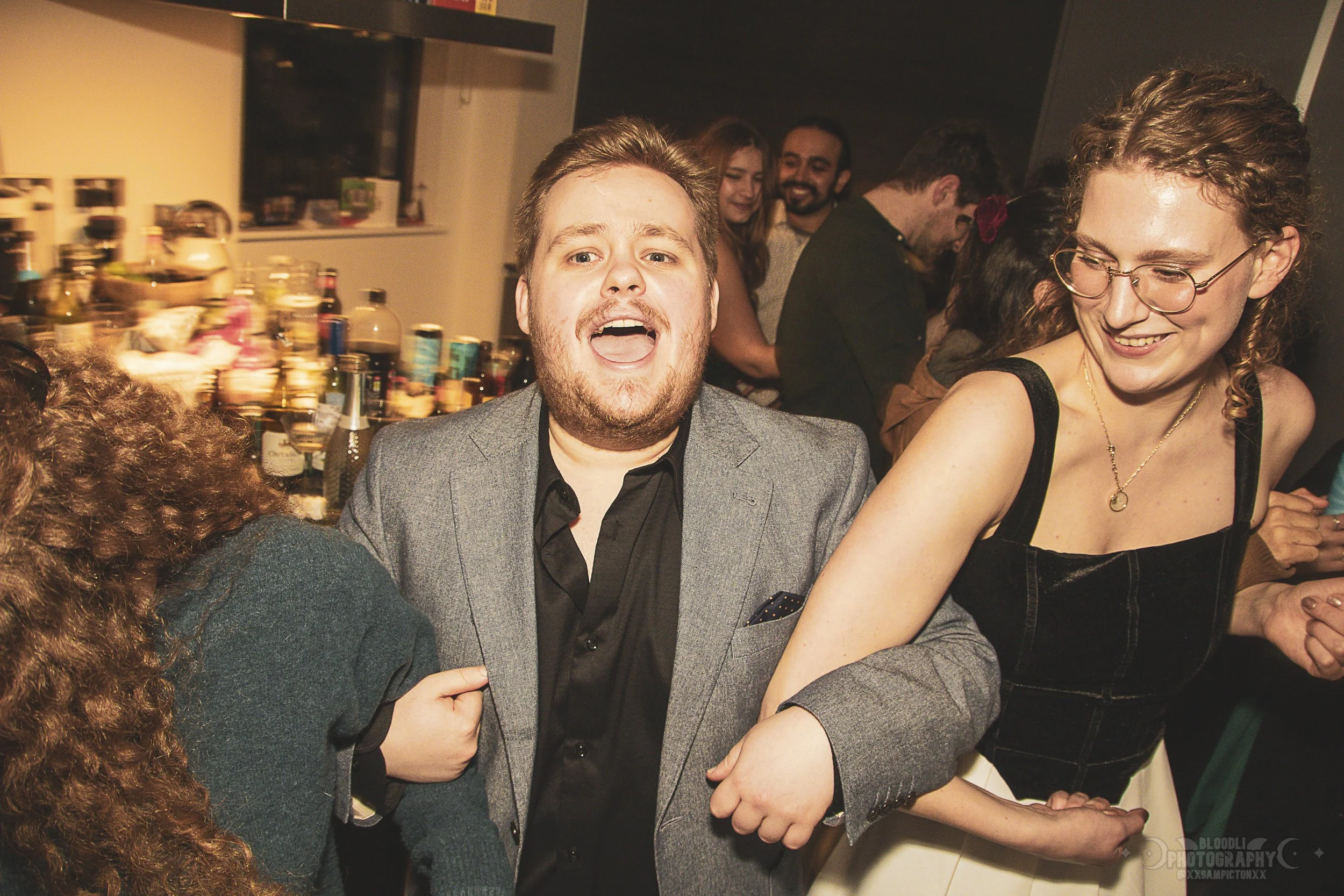 People dancing and enjoying a party in a crowded indoor setting, with some bar supplies visible in the background.