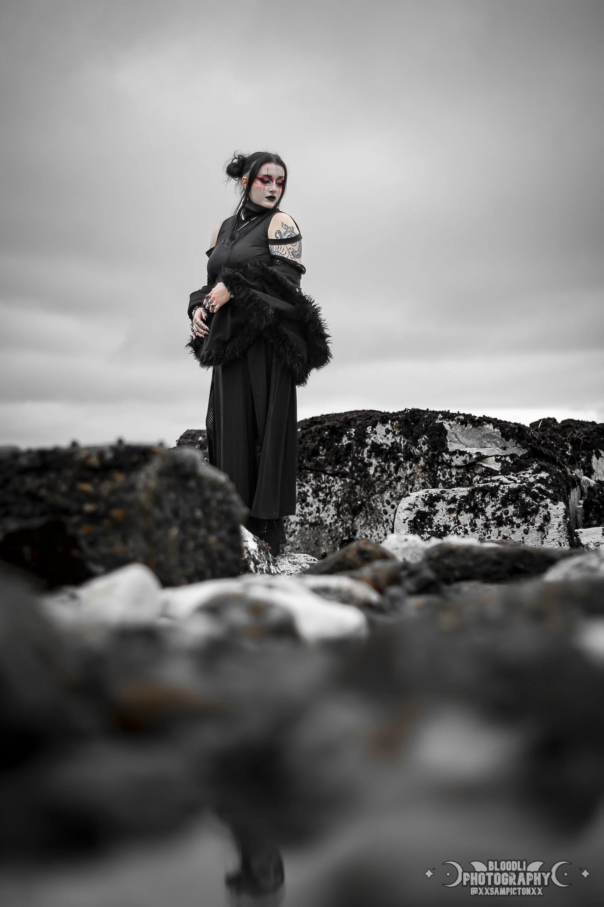 A woman in black gothic fashion standing on rocks by the water, with tattoos and dark makeup, under a cloudy sky.