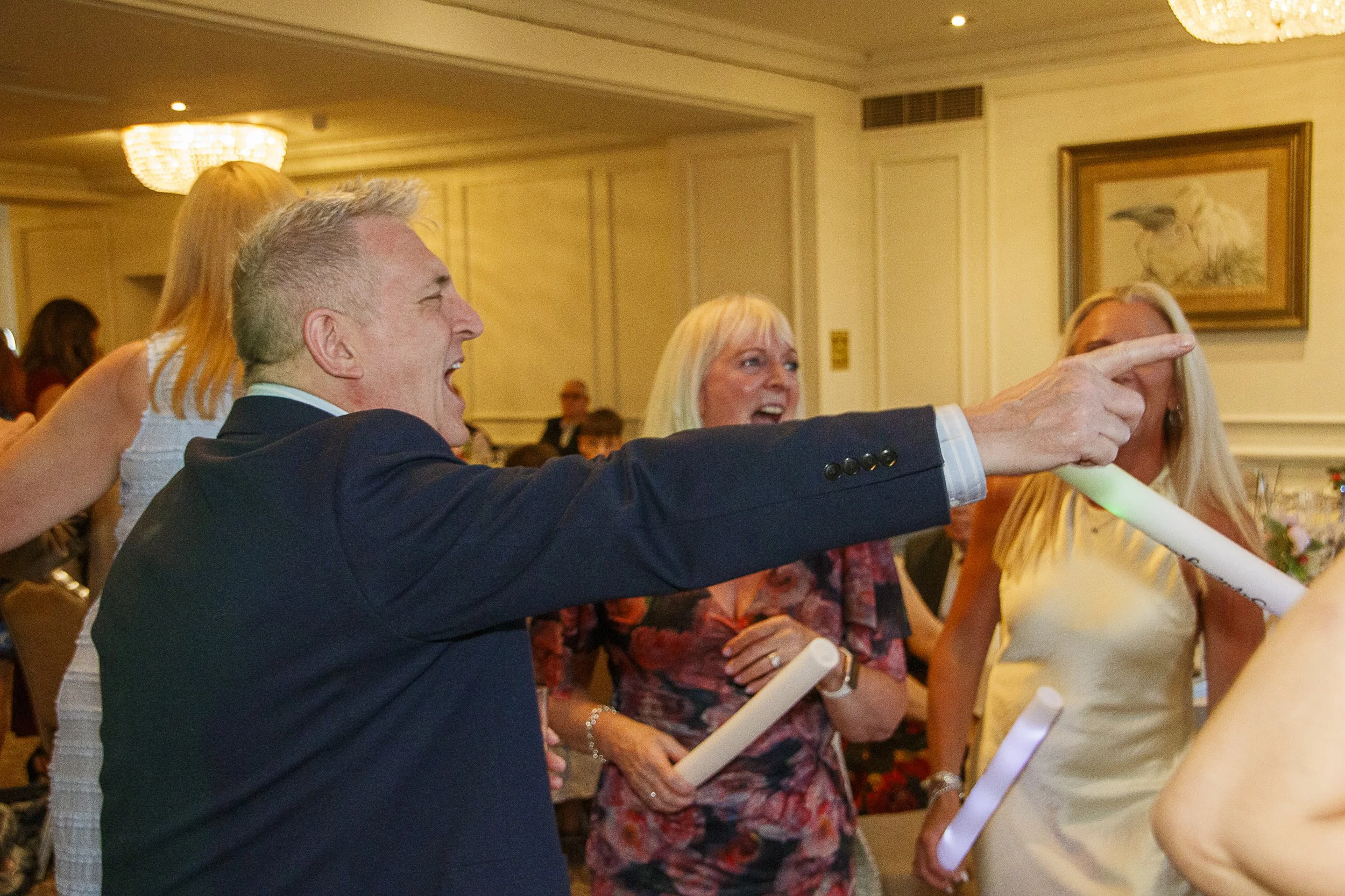 People at a celebration, with one man in a suit enthusiastically pointing and others smiling, holding paper scrolls, in a decorated banquet hall with artwork on the walls.