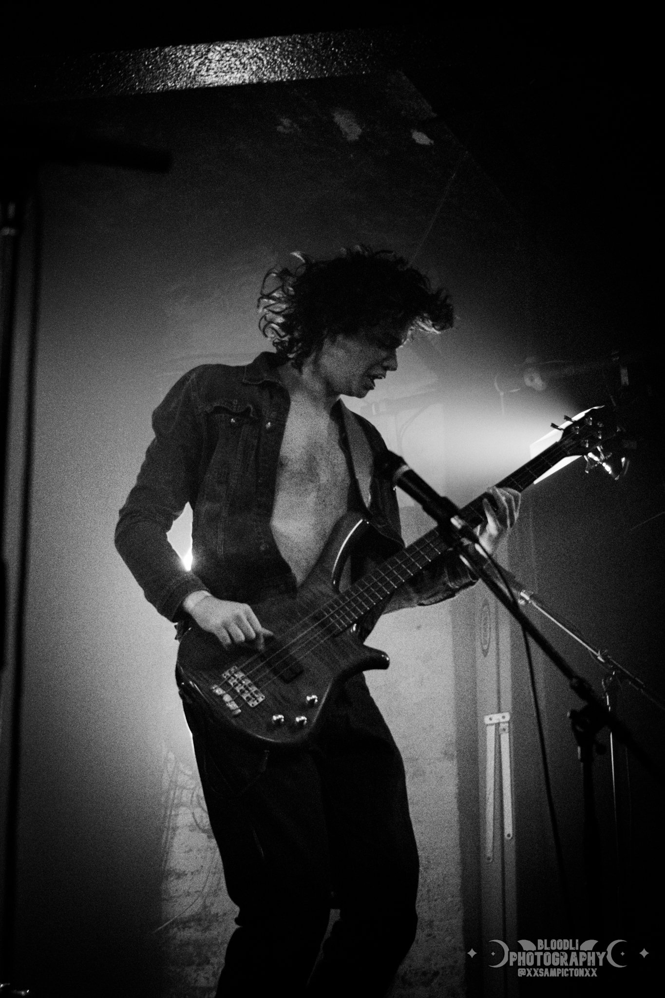 A black and white photo of a male musician playing an electric bass guitar on stage, with messy curly hair and an open shirt
