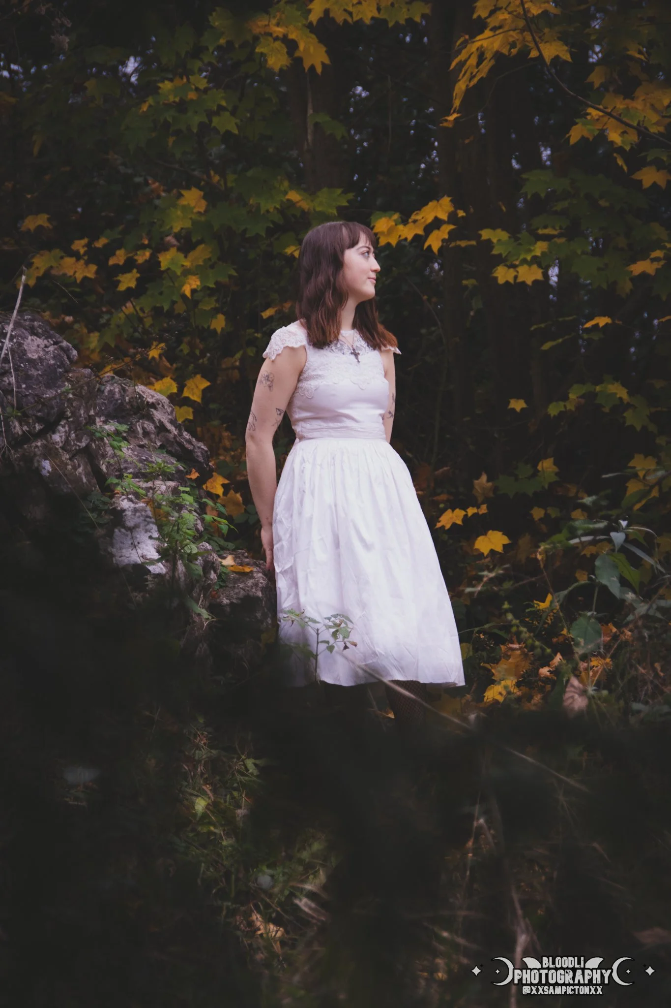 A woman in a white dress standing outdoors in a wooded area with yellow and green leaves. She is looking to the right, with tattoos on her arms and shoulder, and her hair is shoulder-length and brown.