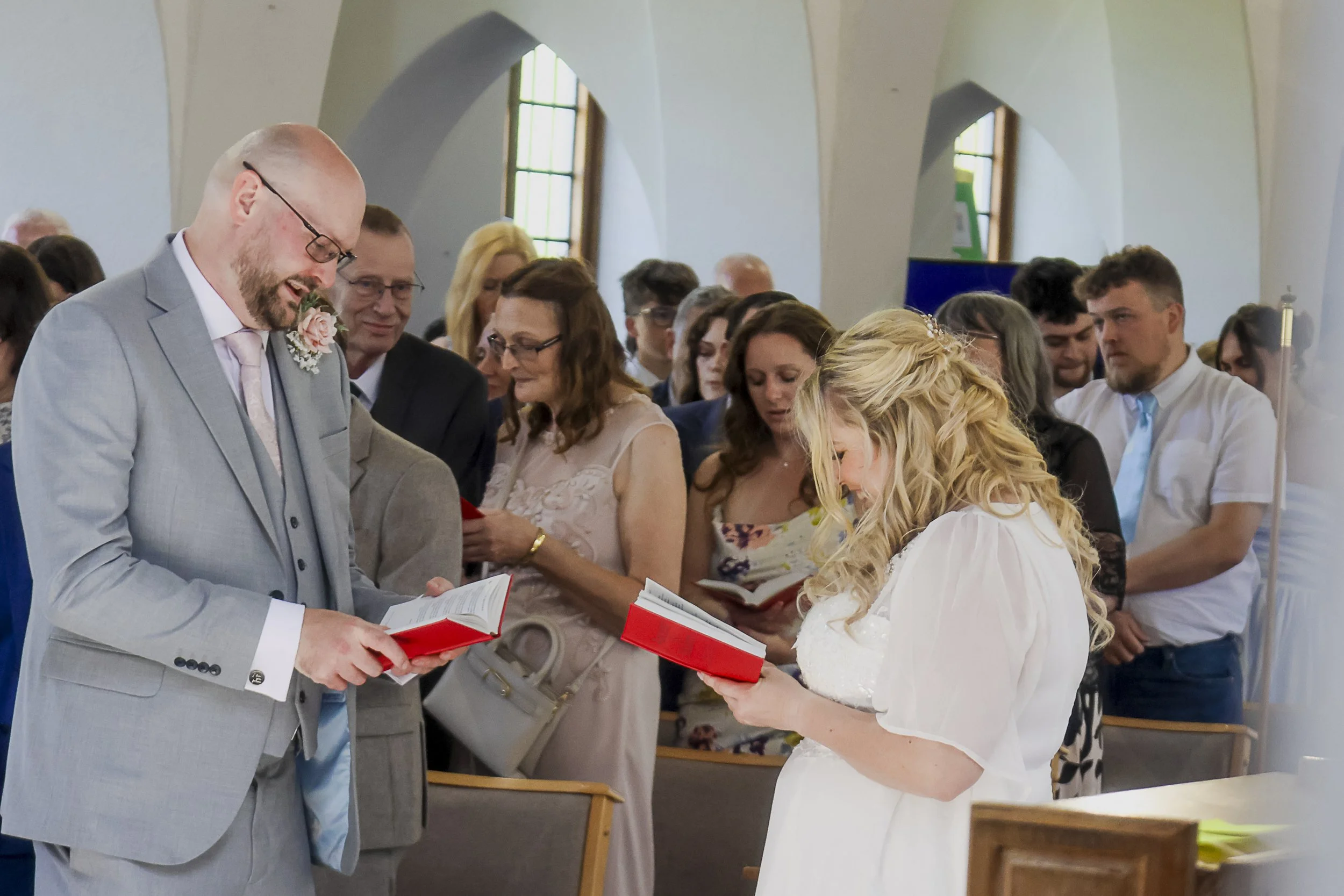 Wedding ceremony with a bride in a white dress and a groom in a light gray suit holding red hymn books, surrounded by guests indoors, some reading from hymn books, in a church with arched windows.