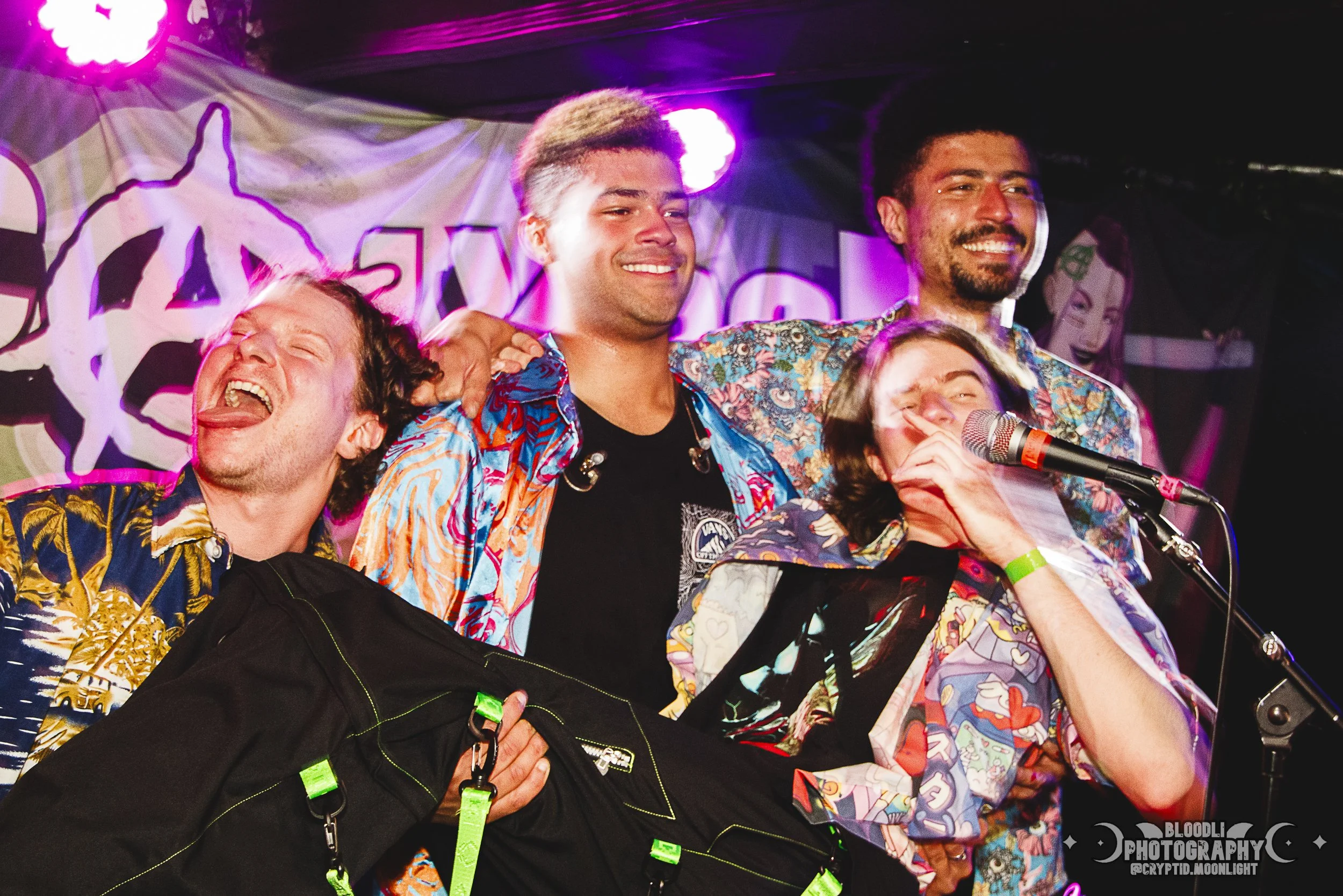 Group of five people on stage, celebrating or performing, wearing colorful Hawaiian shirts, with a vibrant banner in the background, stage lights shining, one person holding a bag, and another speaking into a microphone.