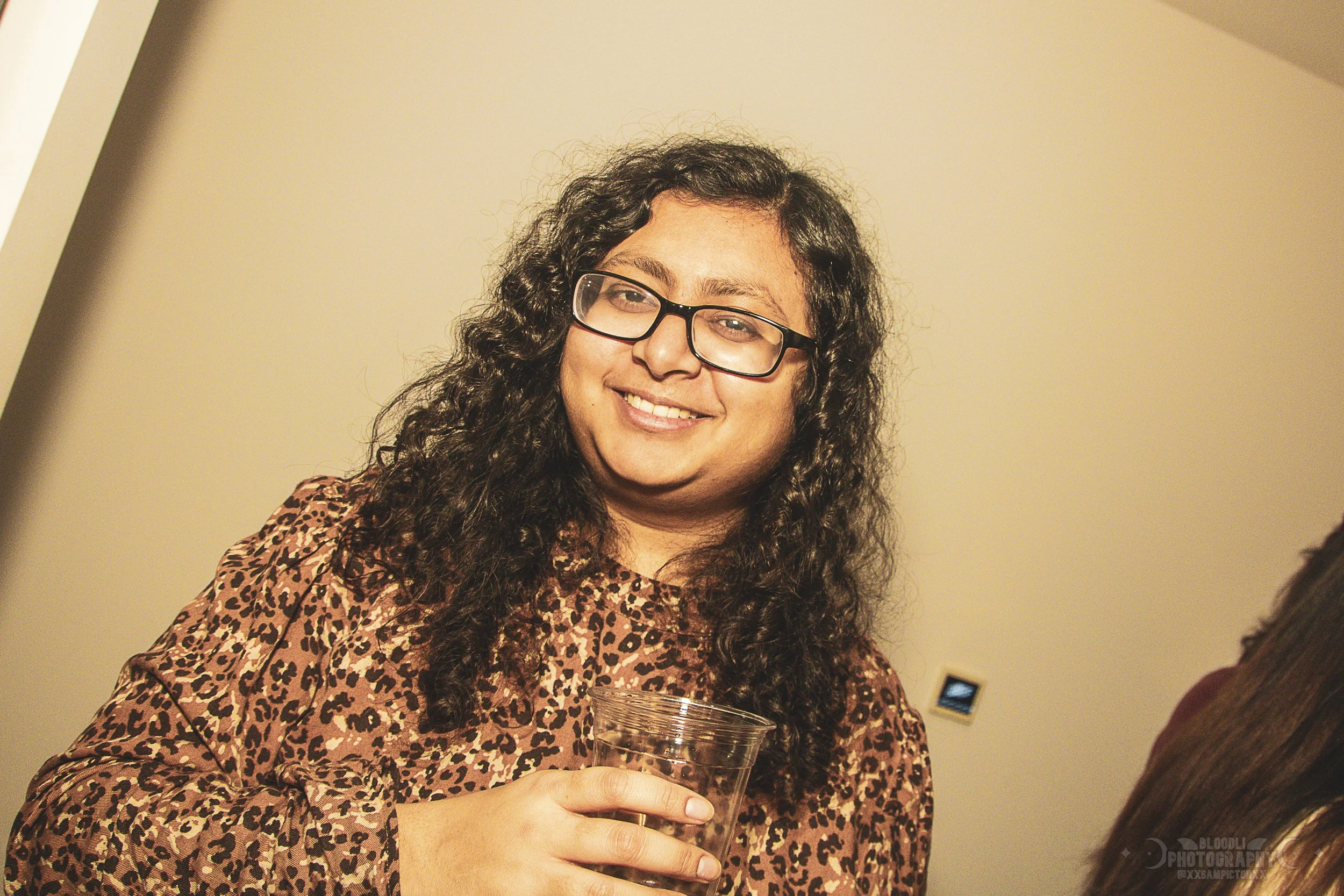 A woman with curly hair, glasses, and a leopard print top holding a clear plastic cup, smiling, indoors.
