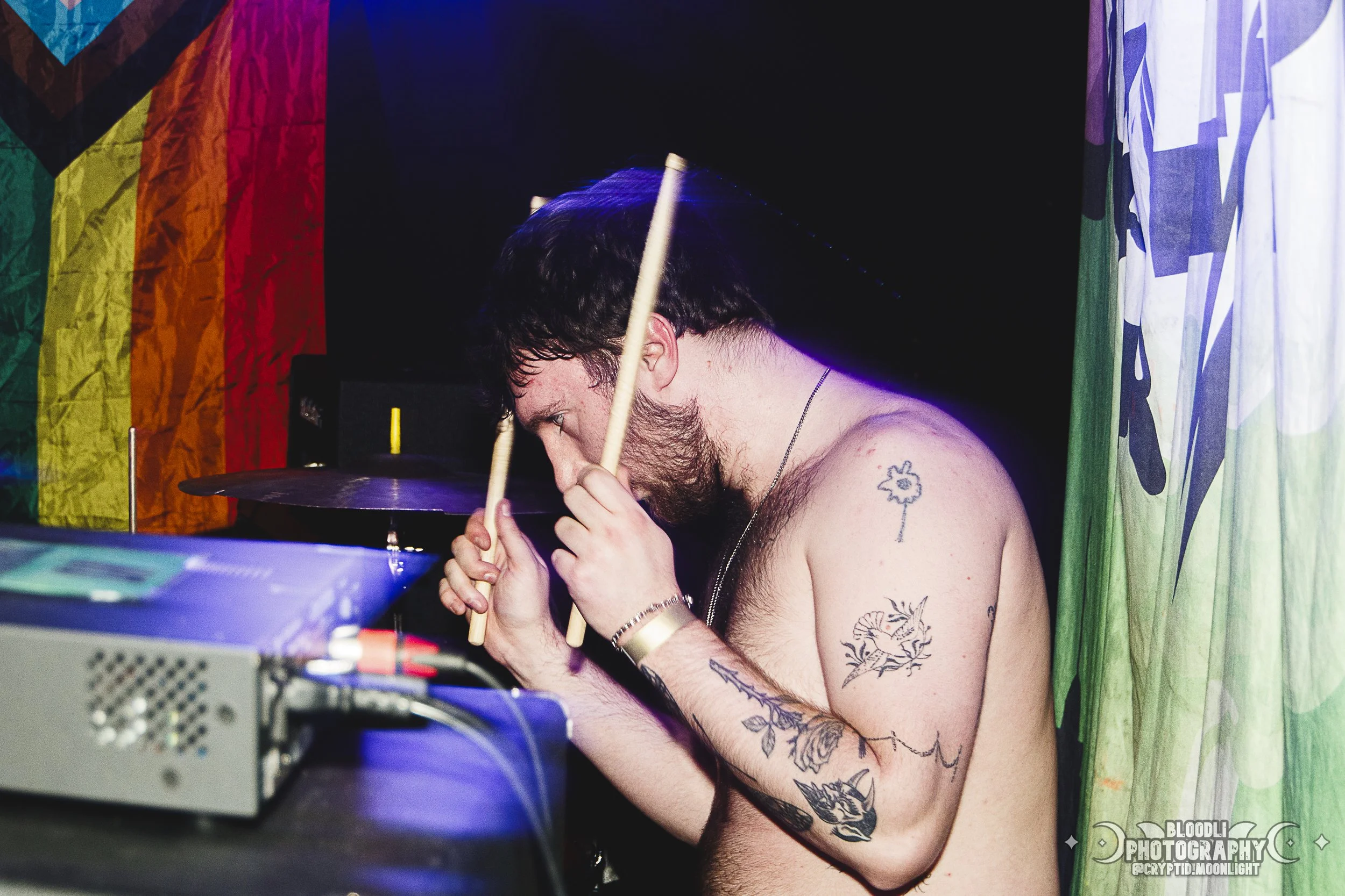 A shirtless man with dark hair and tattoos on his arm and shoulder, holding drumsticks near his face in front of a drum set in a colorful room.