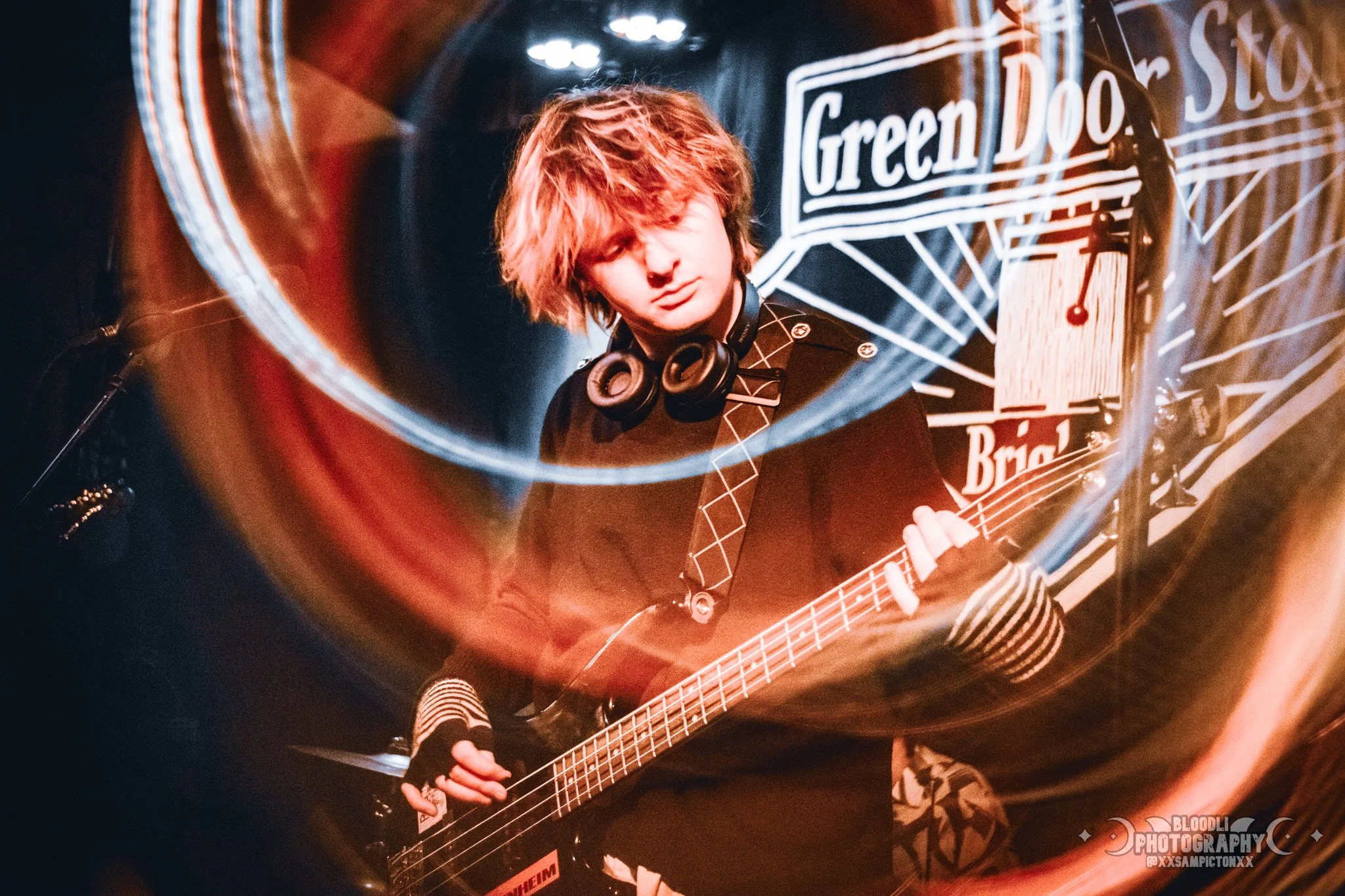 A young person with messy, light brown hair wearing headphones around their neck, playing a bass guitar during a performance. There is a dark background with a banner that reads 'Green Door Studio' and visible stage lighting effects.