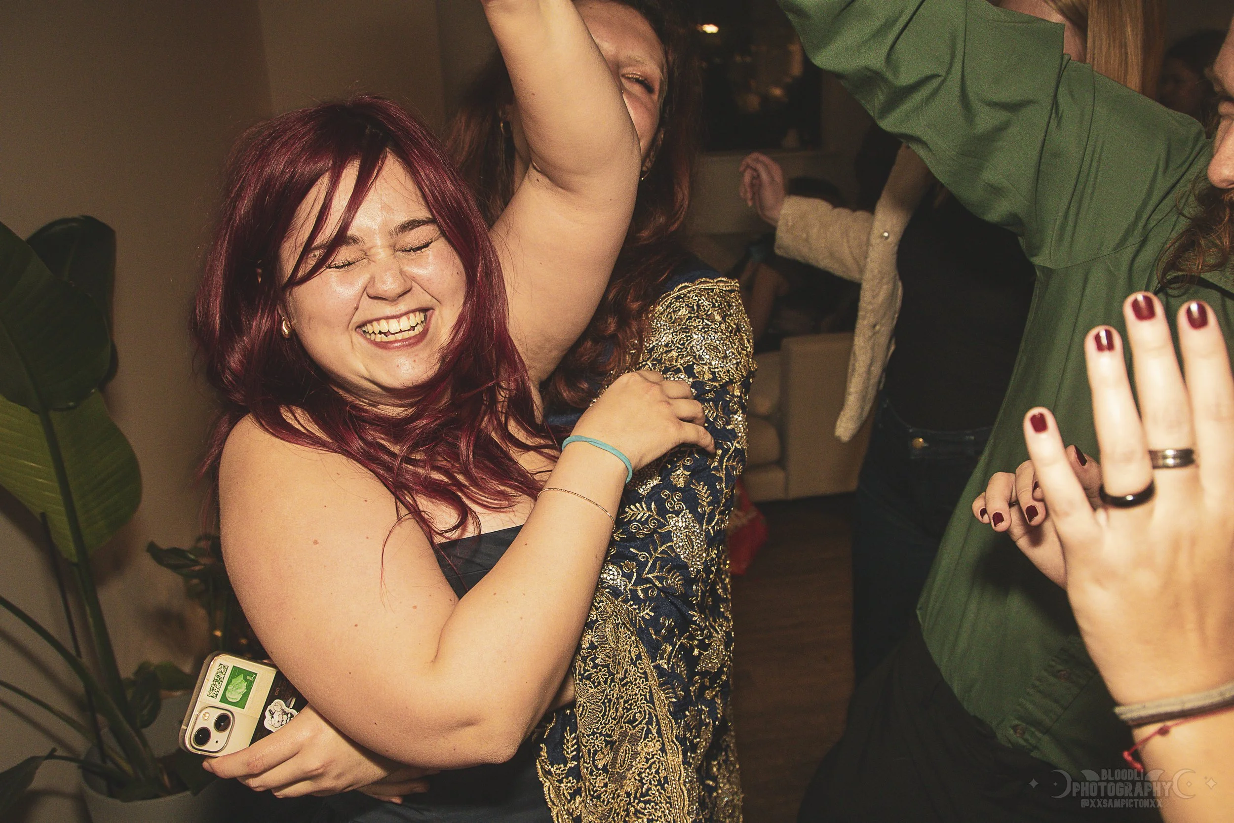 Women dancing and smiling at a party, with one woman wearing a black dress and another in an ornate gold and blue outfit.