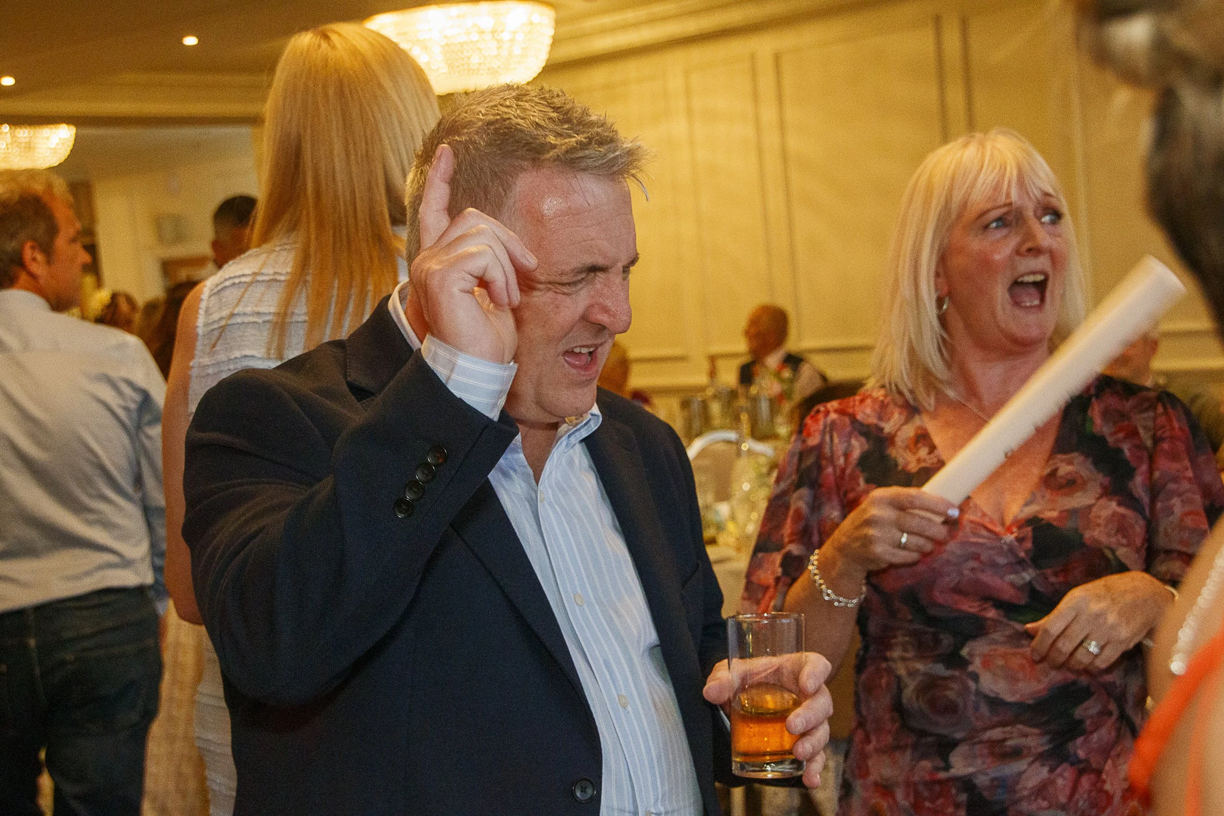 A man in a black suit holding a glass of beer, talking and smiling at a woman with blonde hair wearing a floral dress, in a warmly lit indoor social gathering.