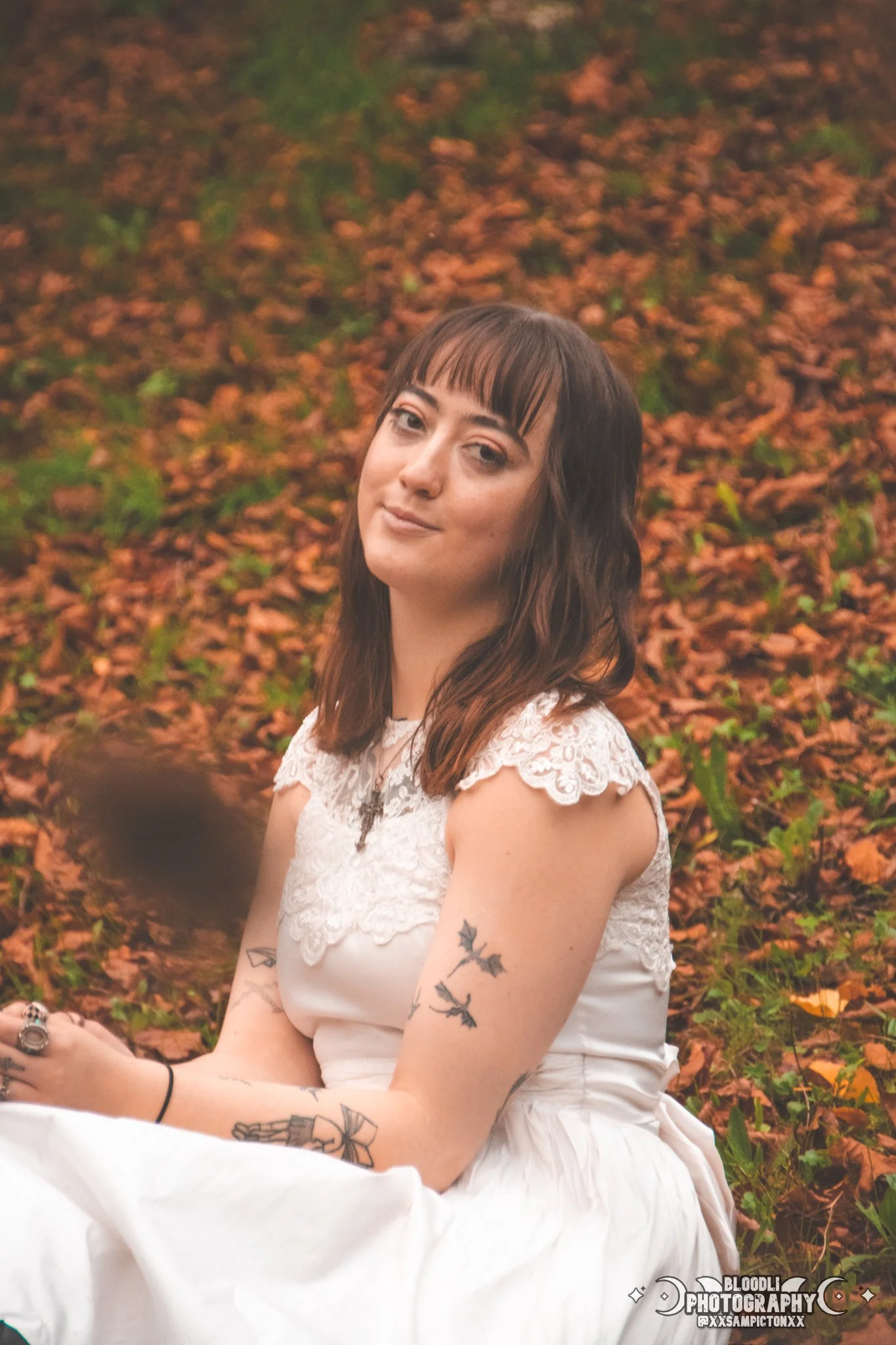 A woman with brown hair and tattoos sitting outdoors on the ground covered in orange and green leaves, wearing a white dress with lace details, looking at the camera with a slight smile.
