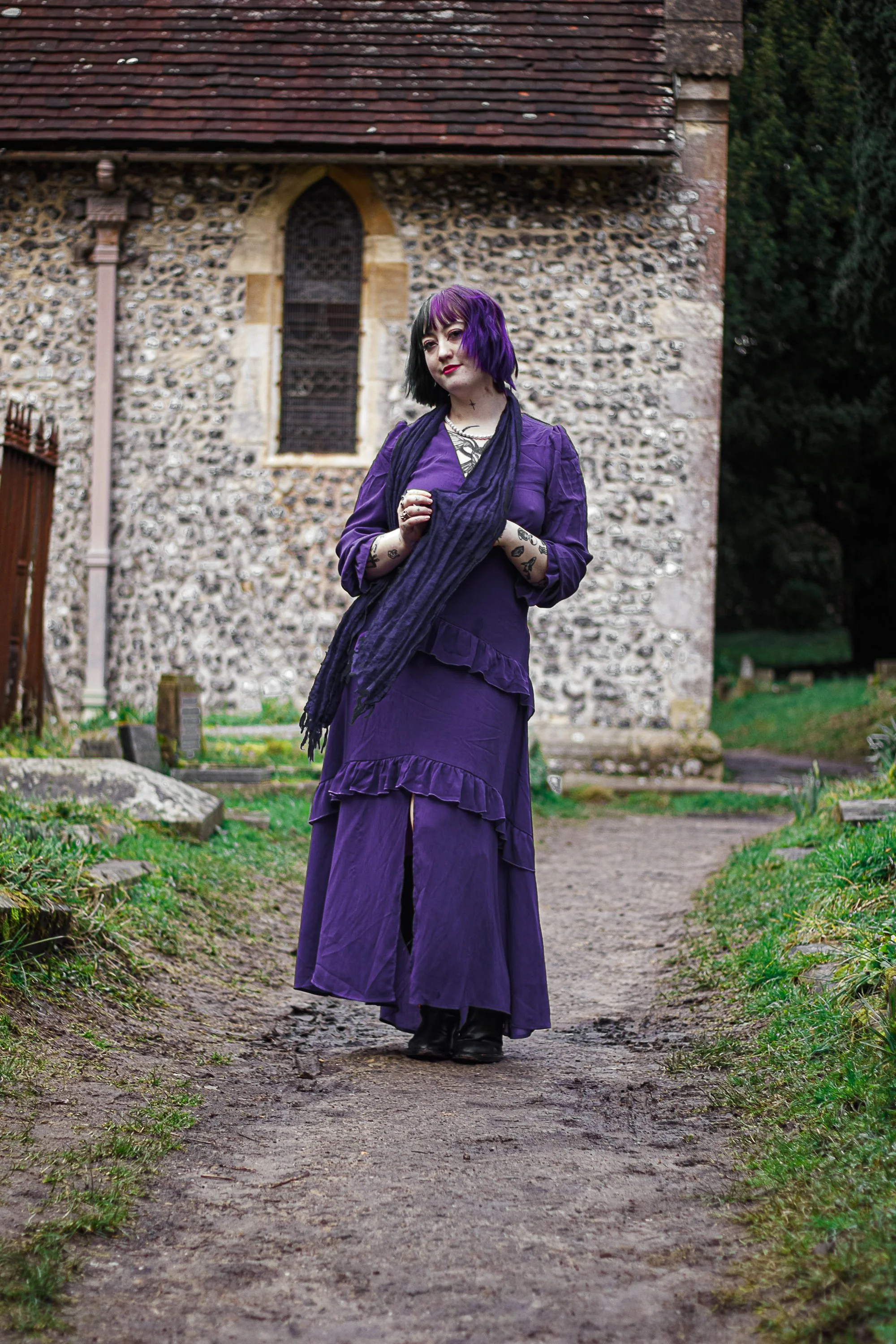 A young woman with black and purple hair standing on a dirt path in front of an old stone church, wearing a purple dress, black boots, and a dark scarf, with tattoos visible on her arms.