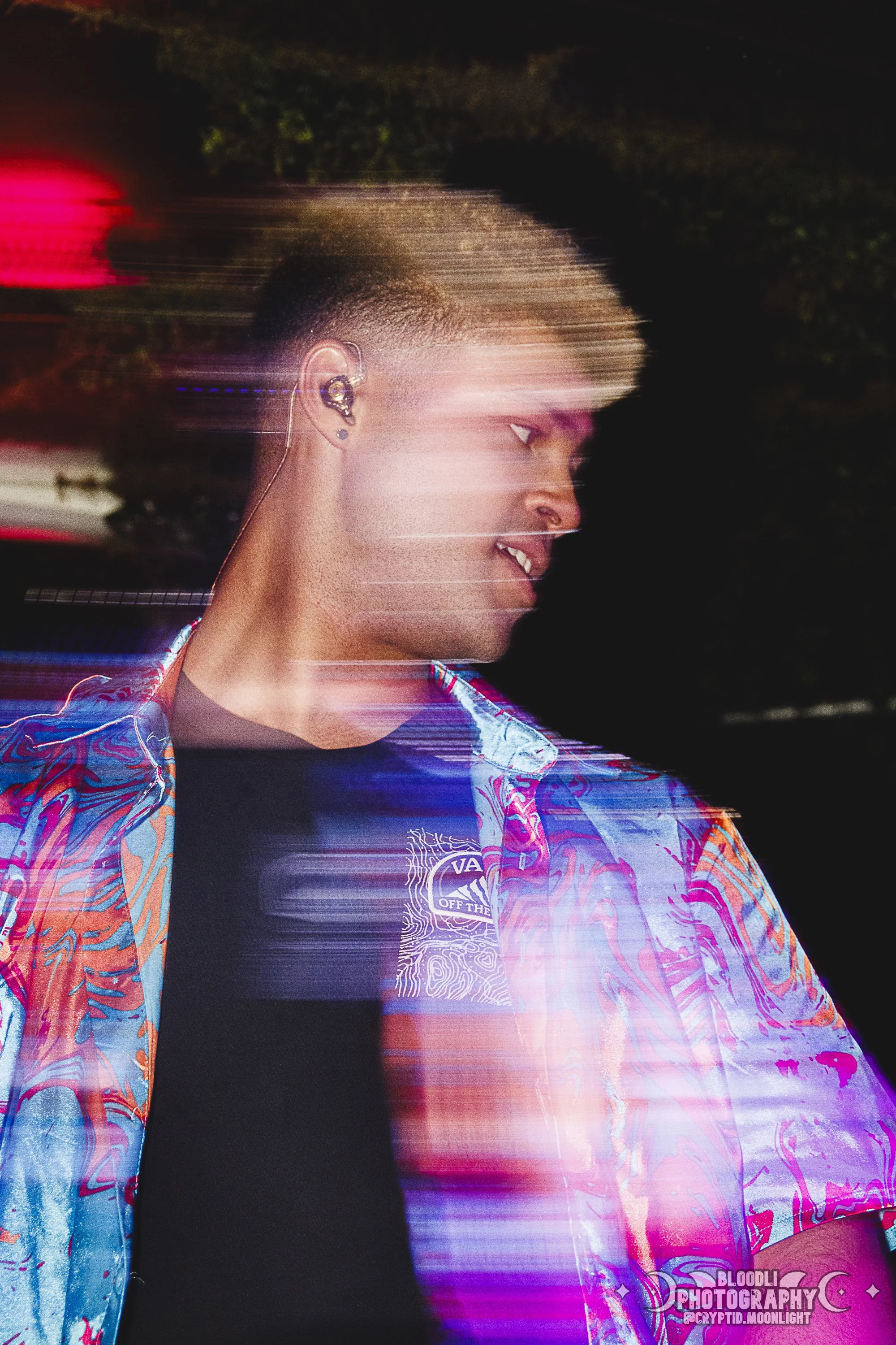 A young man with short hair is smiling and looking to the side, wearing an earpiece and a colorful patterned shirt at night, with light trails and motion blur effect.