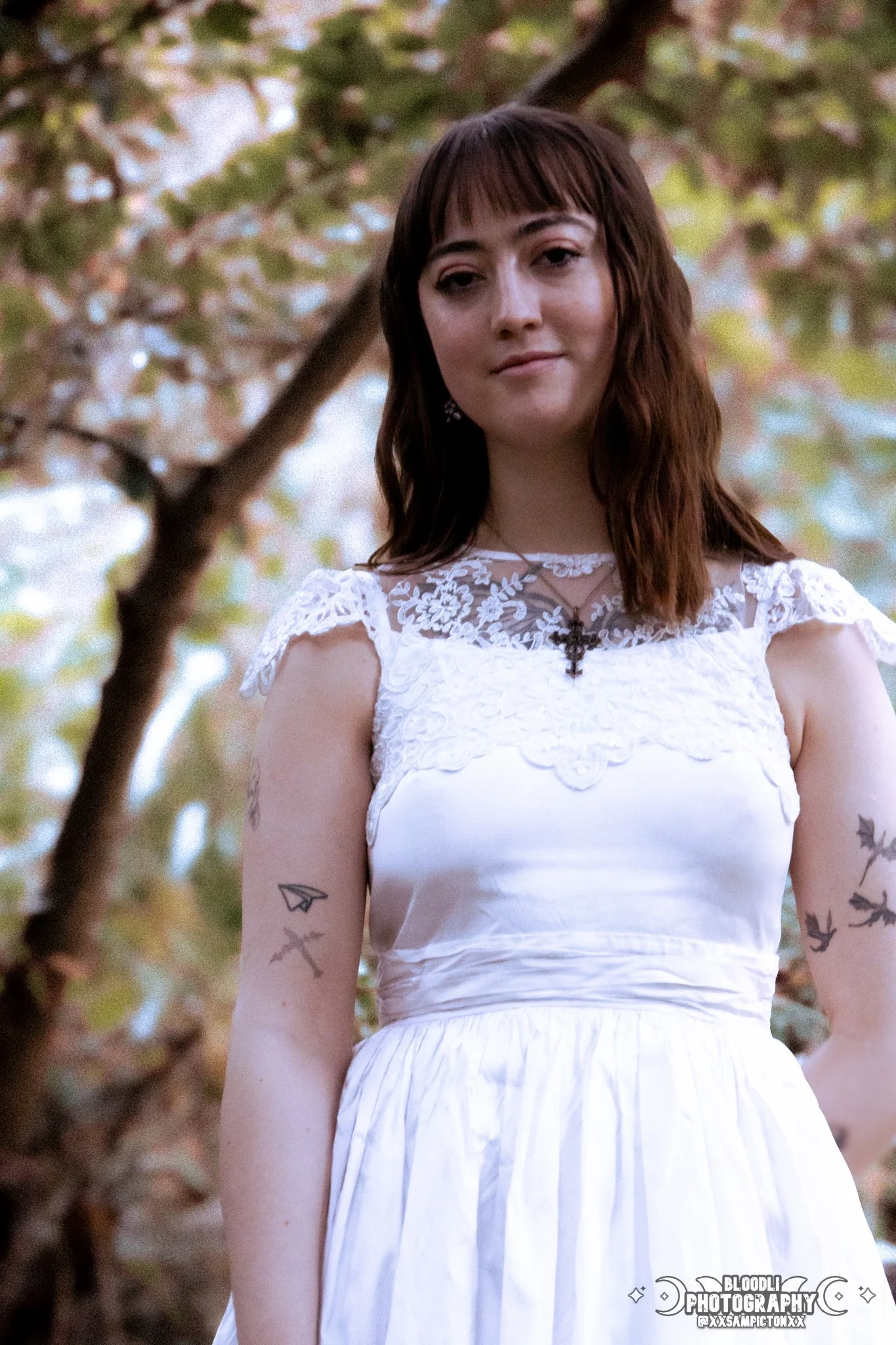 A young woman with brown hair and tattoos on her arms, wearing a white lace dress and a necklace with a cross, standing outdoors with trees and leaves in the background.