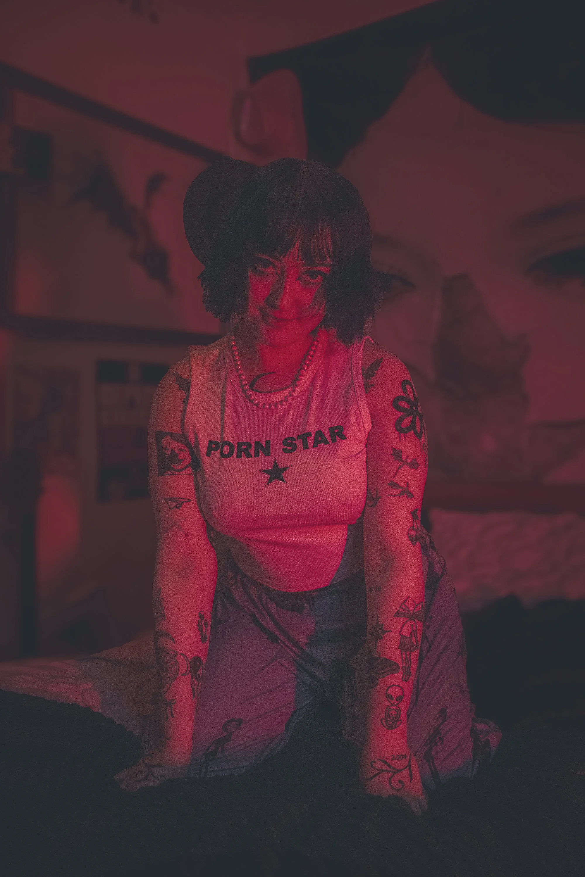 A person with shoulder-length dark hair, tattoos on arms, wearing a white sleeveless shirt that reads 'PORN STAR,' a beaded necklace, and patterned pants, is kneeling on a bed in a dimly lit room with red lighting.