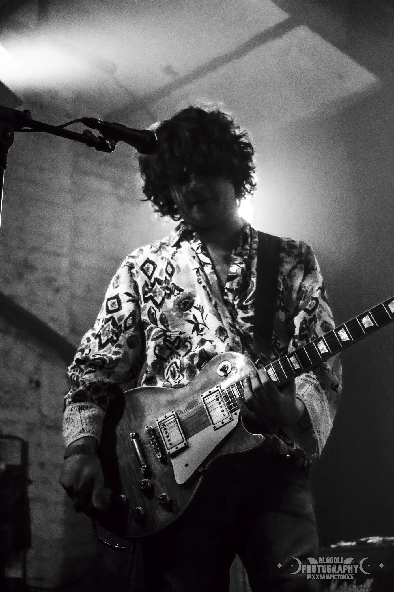A musician with curly hair playing an electric guitar during a performance in a dimly lit setting, captured in black and white.