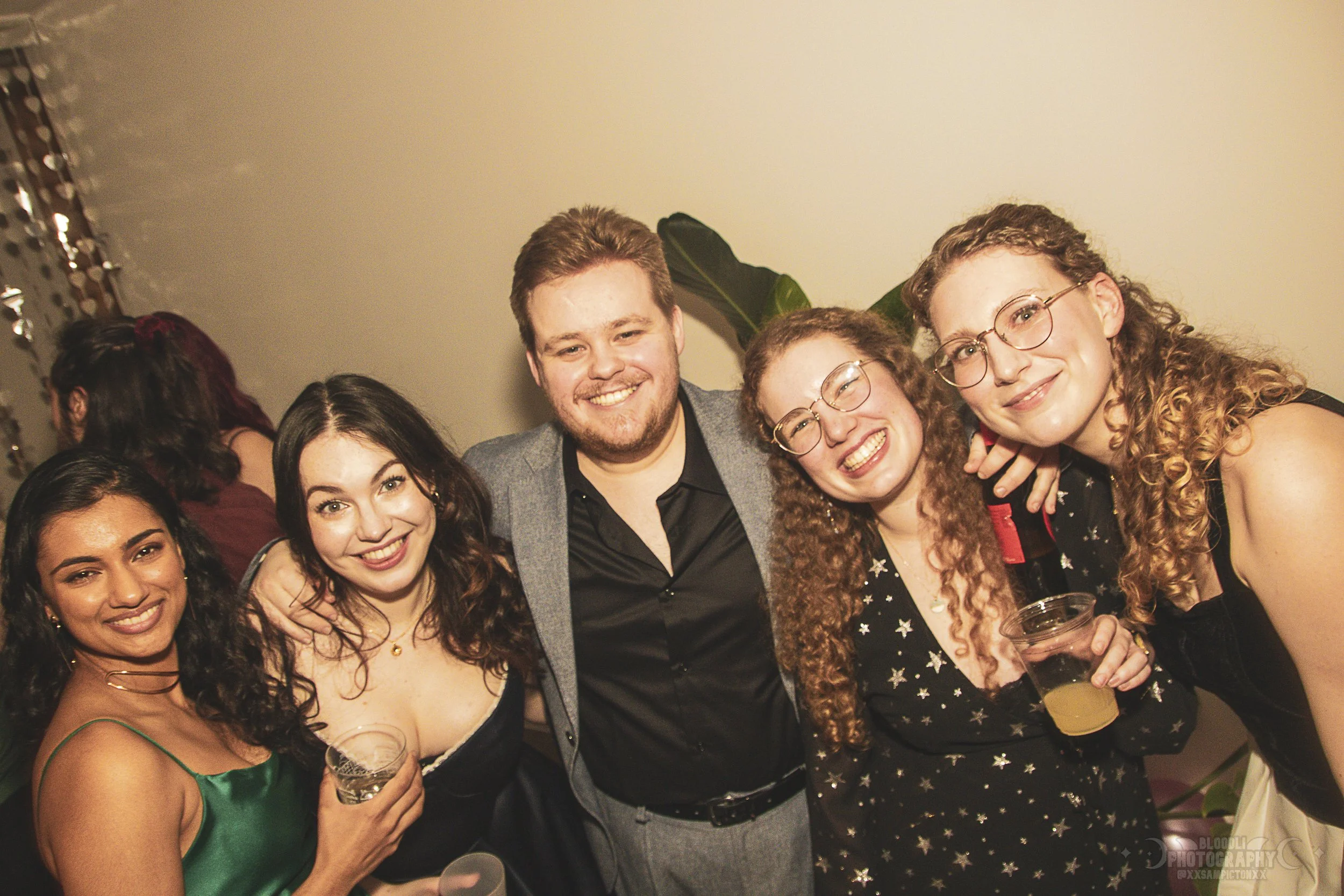 Group of five young adults smiling and posing together at an indoor party, some holding drinks.