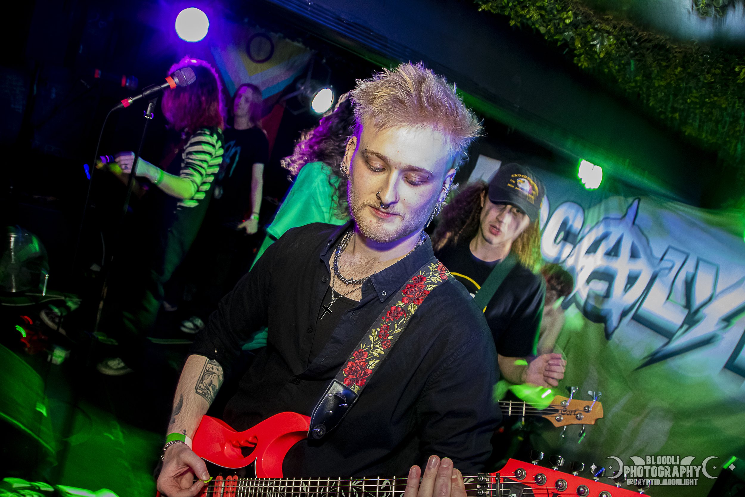 Young man with dyed blonde hair playing red electric guitar, wearing black shirt and floral strap, in a lively music venue with colorful lights and a band performing in the background.