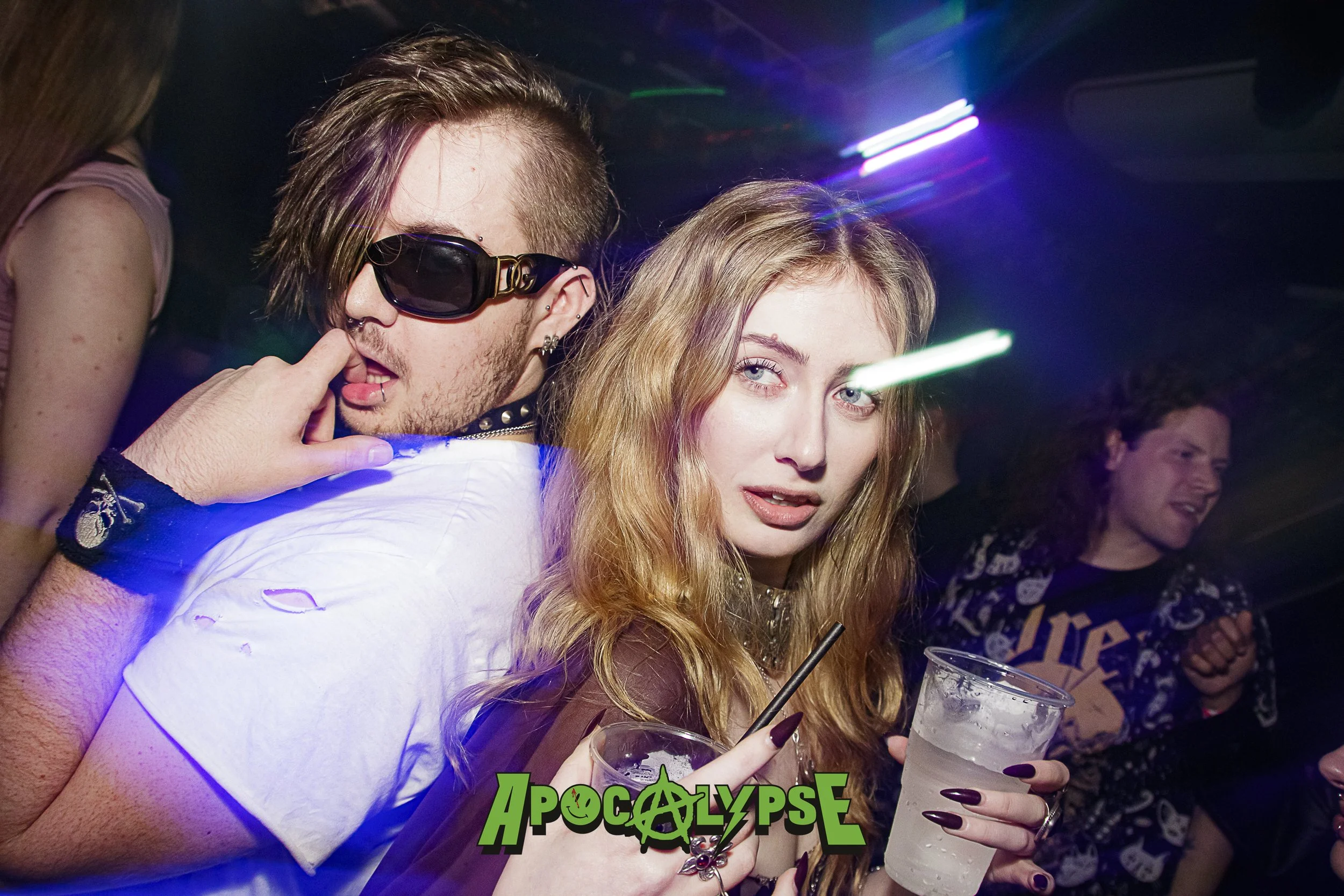 Young woman with long blonde hair holding a drink at a party, with a man with short hair and sunglasses behind her and other party-goers in the background, under colorful lights at a nightclub.